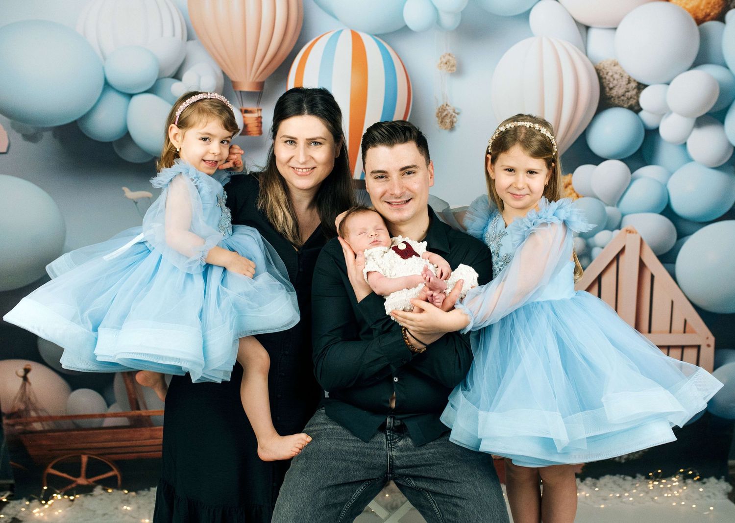 Happy family with two girls in blue dresses and a newborn baby posing in front of colorful balloon backdrop.