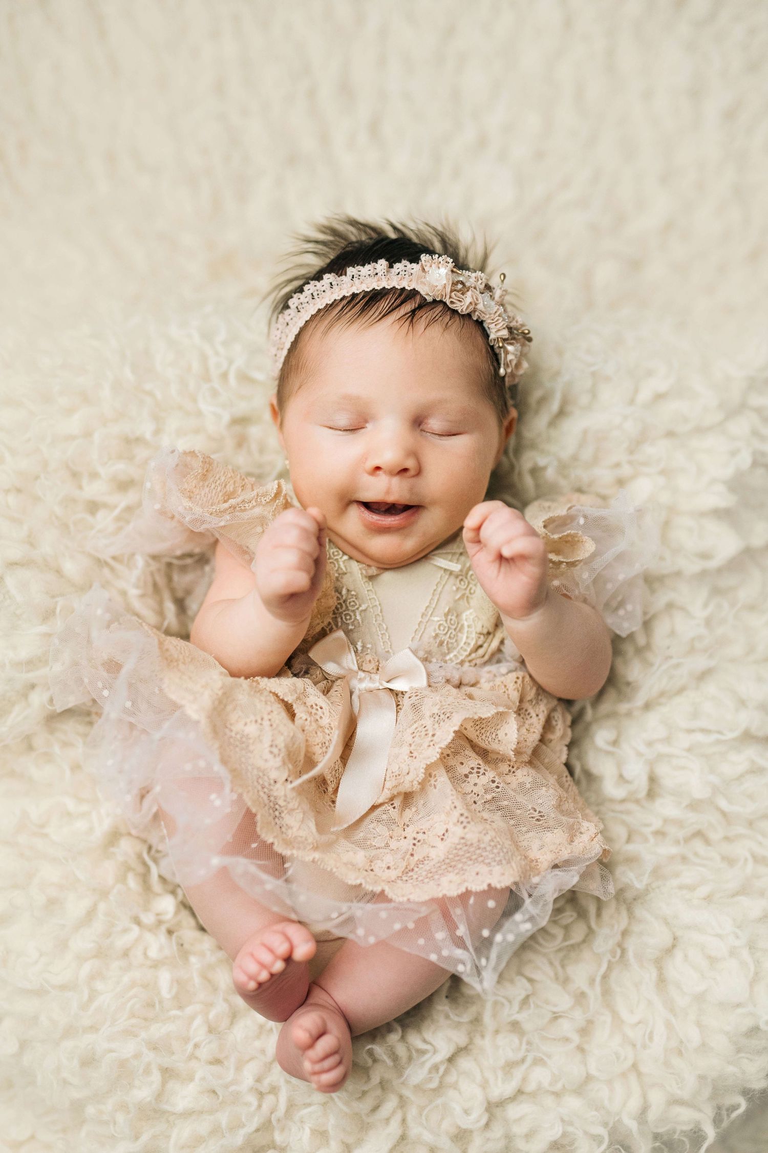 Adorable newborn baby girl smiling in lace dress and floral headband lying on fluffy white blanket.