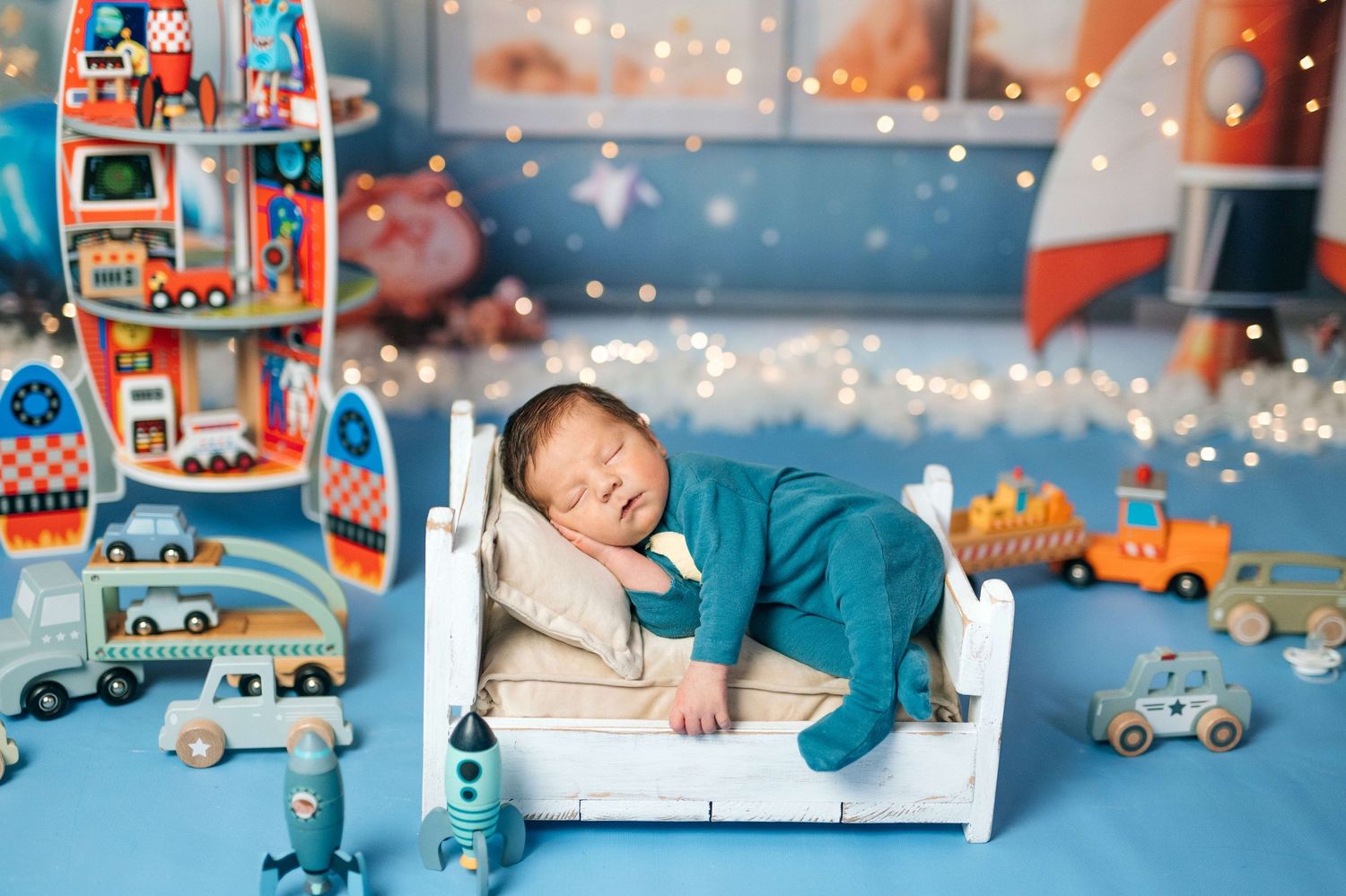 Newborn baby sleeping peacefully on a tiny white bed surrounded by colorful toys and bokeh lights.