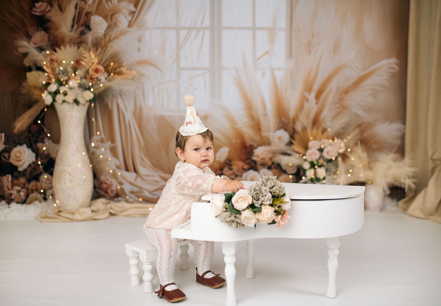 A baby girl in a white dress and party hat sits by a white piano surrounded by flowers for her first birthday.