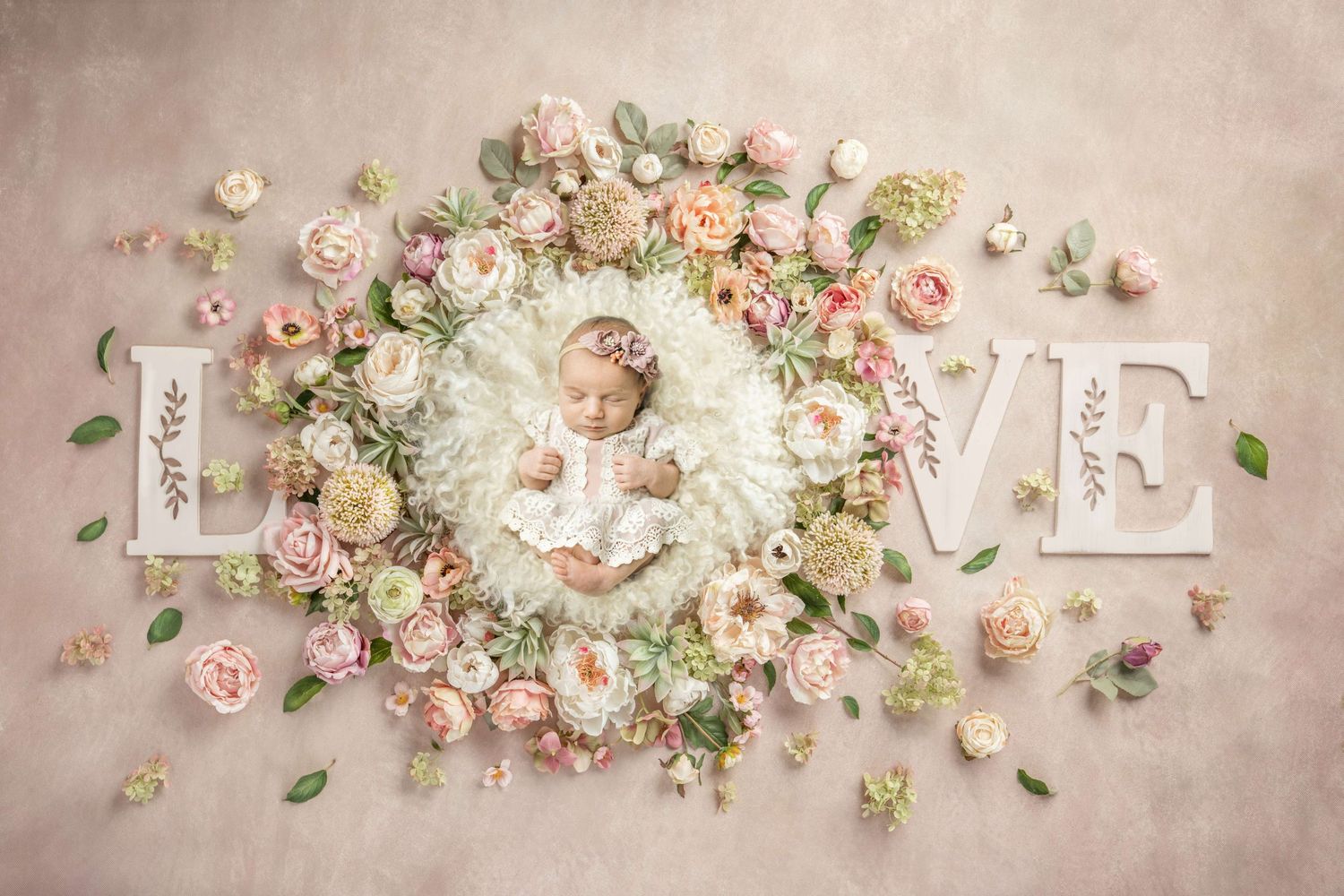 Newborn baby sleeping in floral wreath surrounded by pink roses with decorative LOVE letters on pink background.