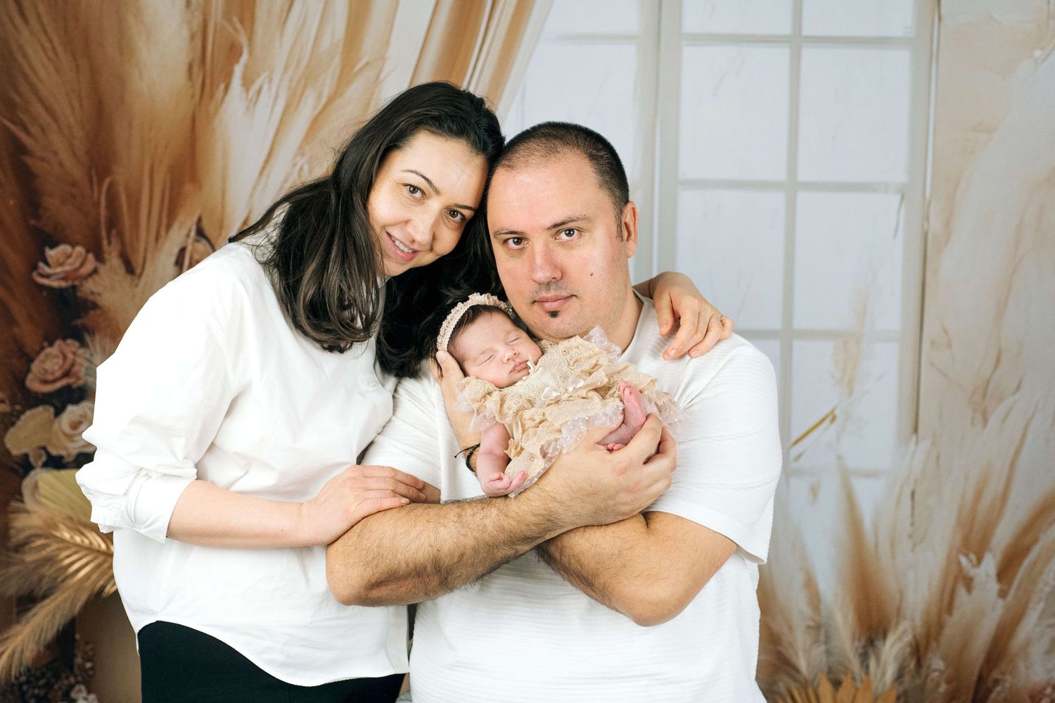 Happy couple in white shirts holding their sleeping newborn baby during a professional studio portrait session.