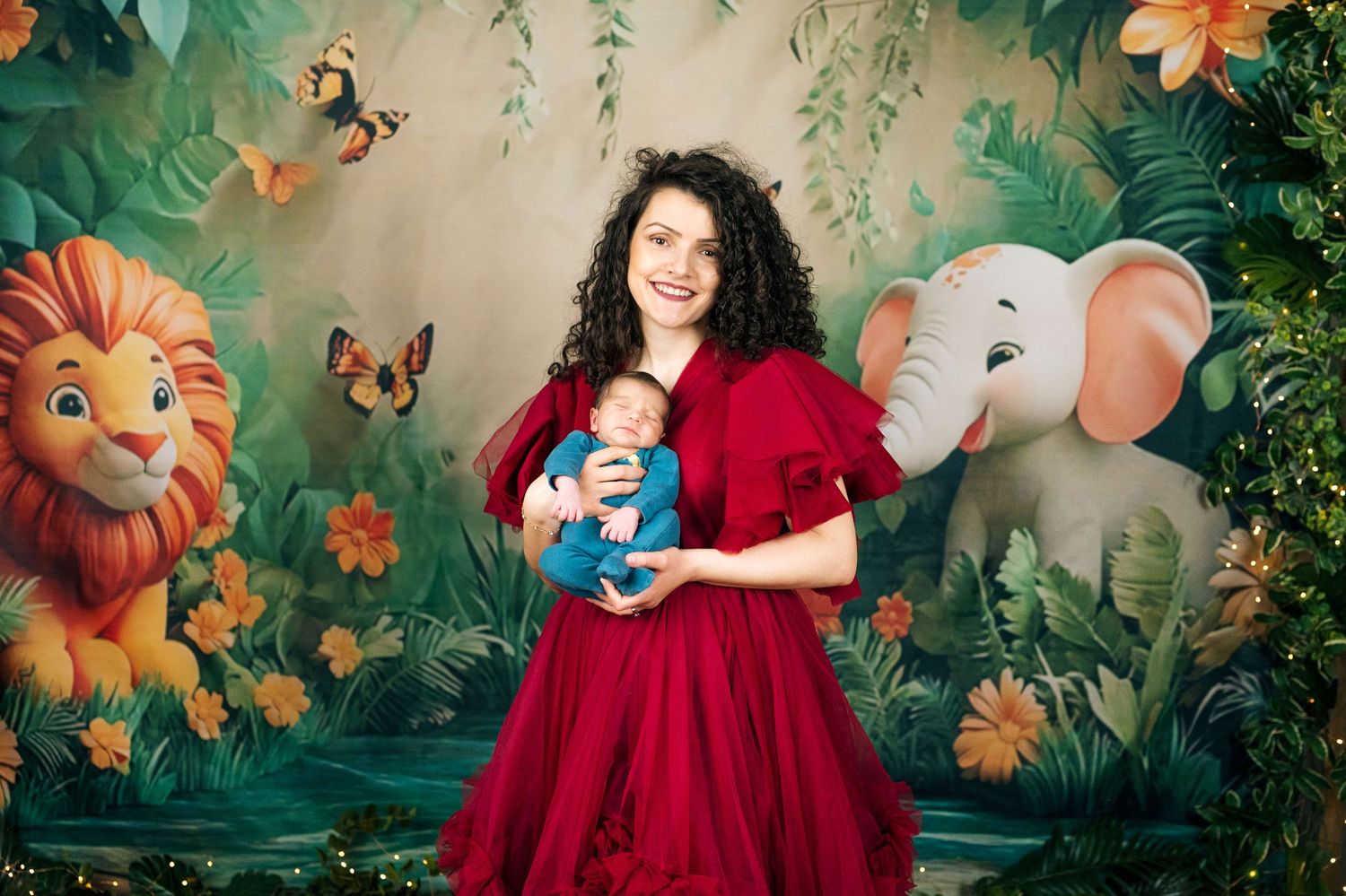 Woman in red dress holding newborn baby in front of jungle-themed backdrop with cartoon lion and elephant.