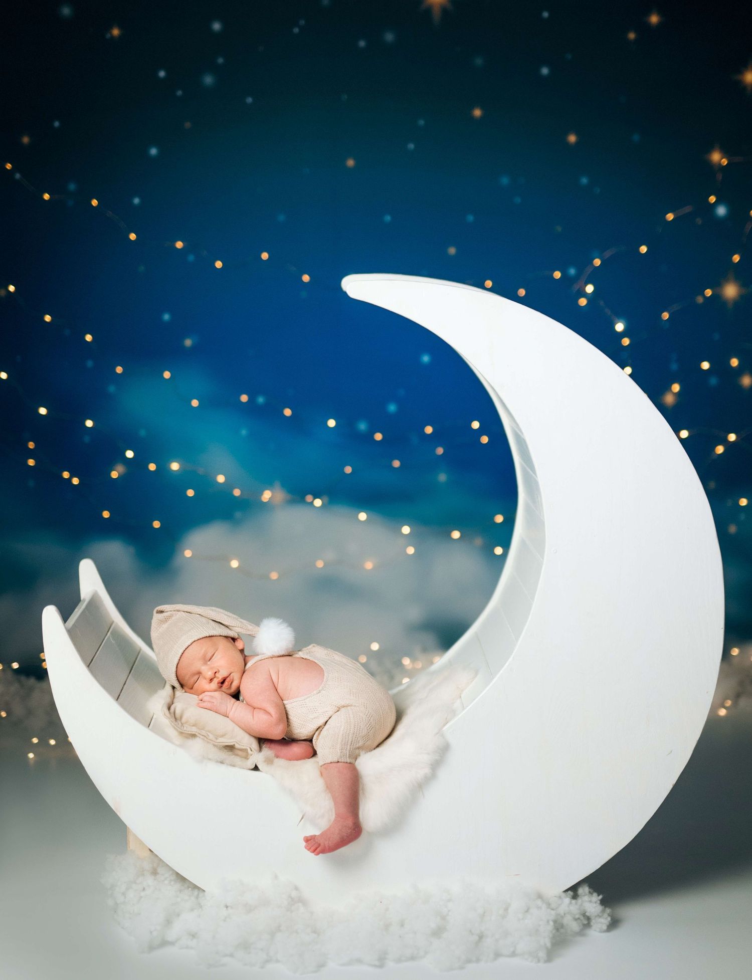 Newborn baby sleeping peacefully on a white crescent moon prop against a dreamy starry night sky backdrop.