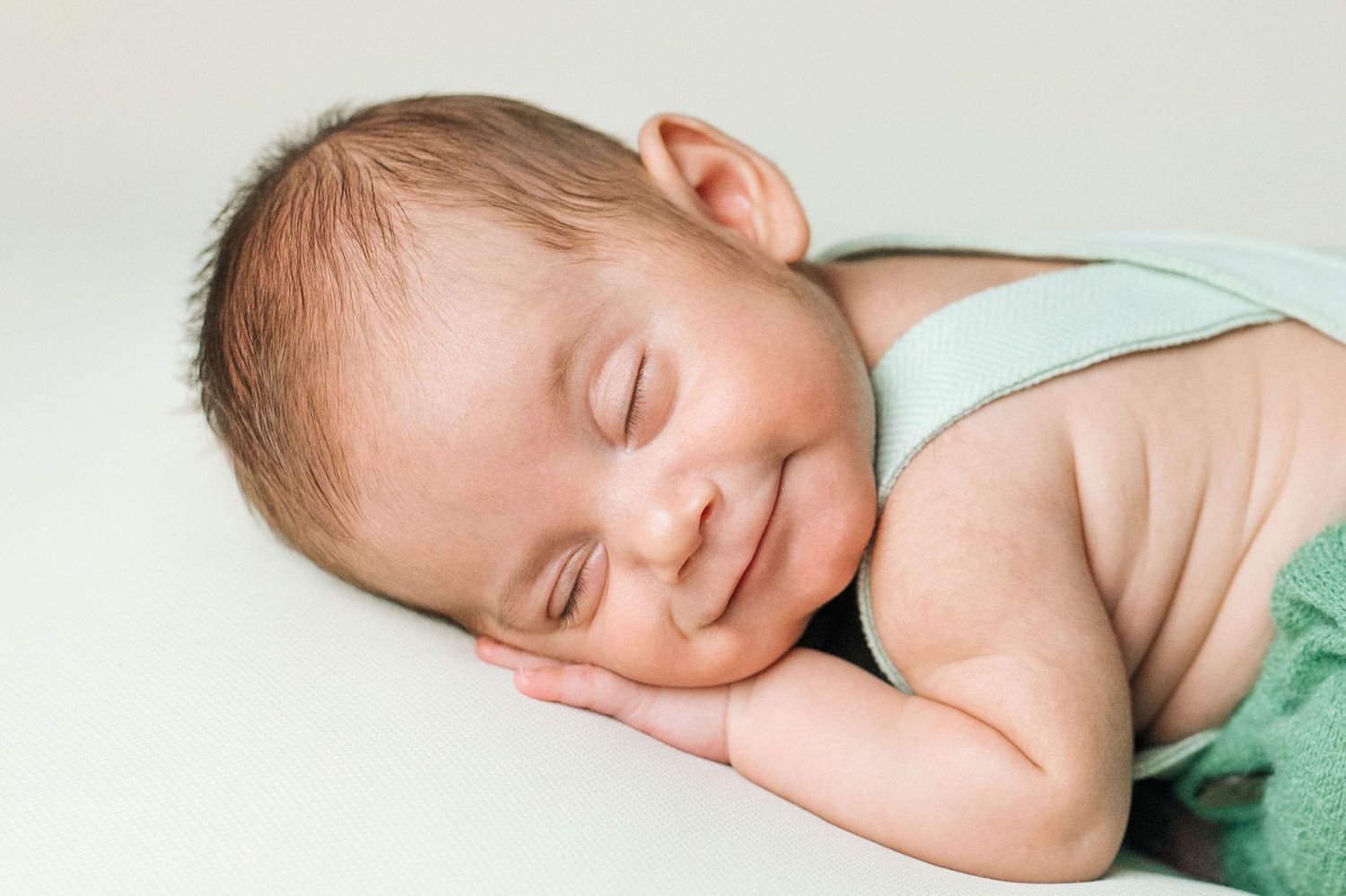 Smiling newborn baby sleeping peacefully on a white background wearing a mint green outfit.