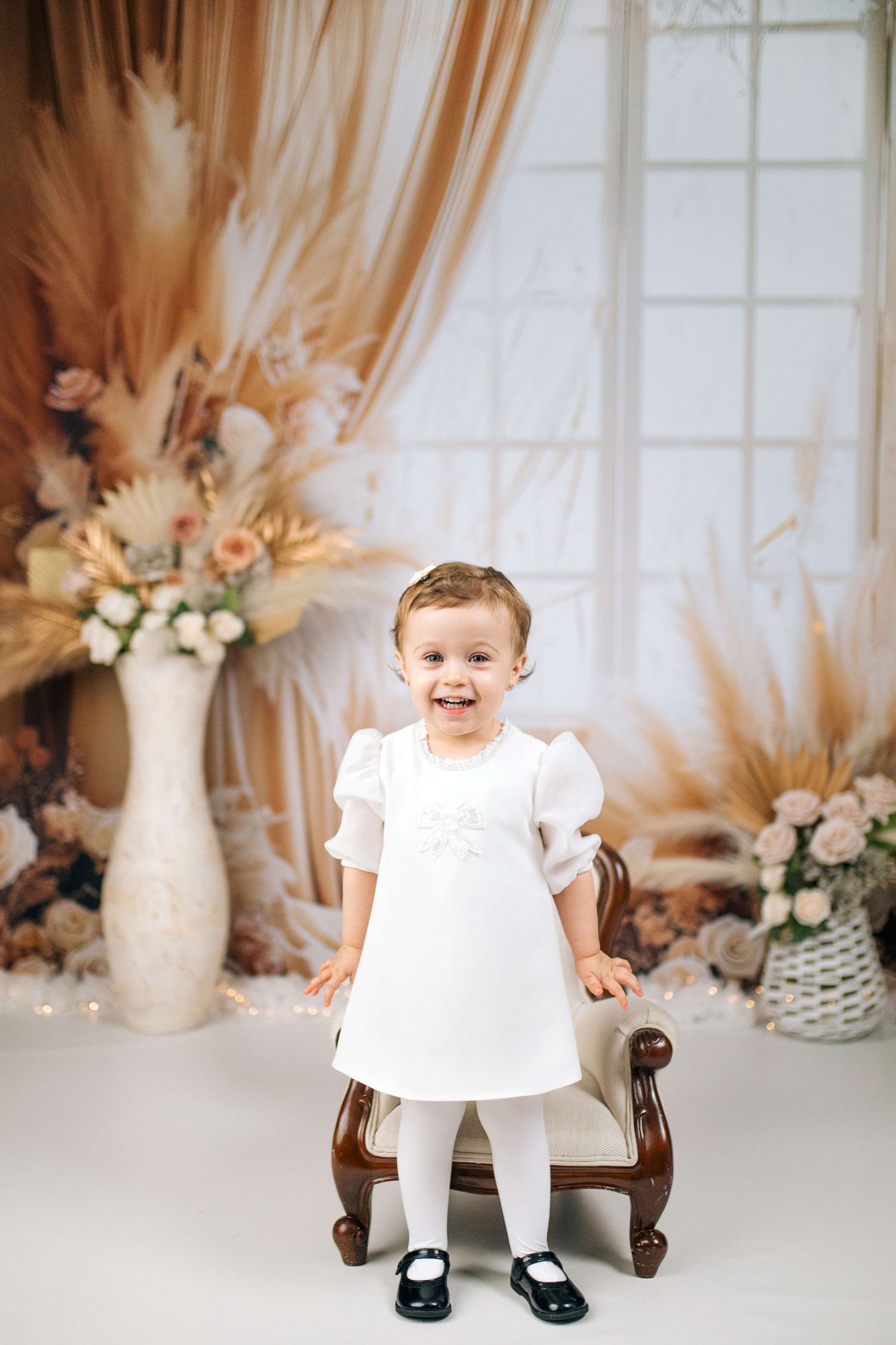 Smiling toddler girl in white dress and black shoes standing in decorated studio with floral backdrop.