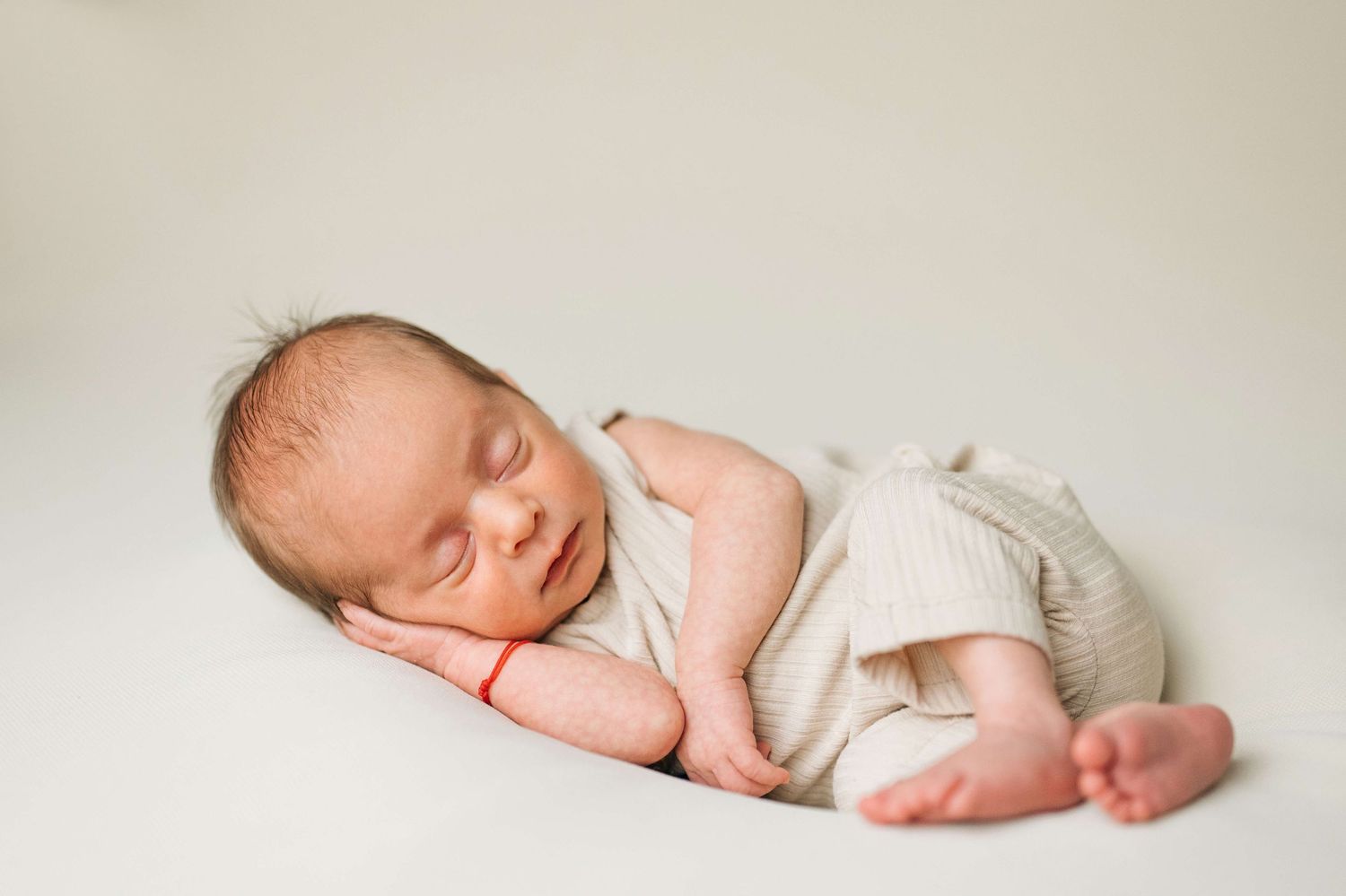 A peaceful newborn baby sleeps curled up in a beige romper on a soft white blanket in a studio photo.