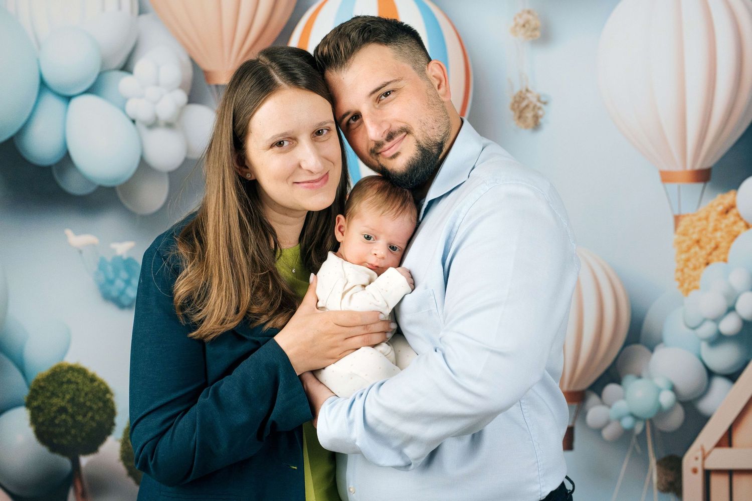 Happy parents smiling and holding their newborn baby in a soft blue balloon-themed studio portrait.