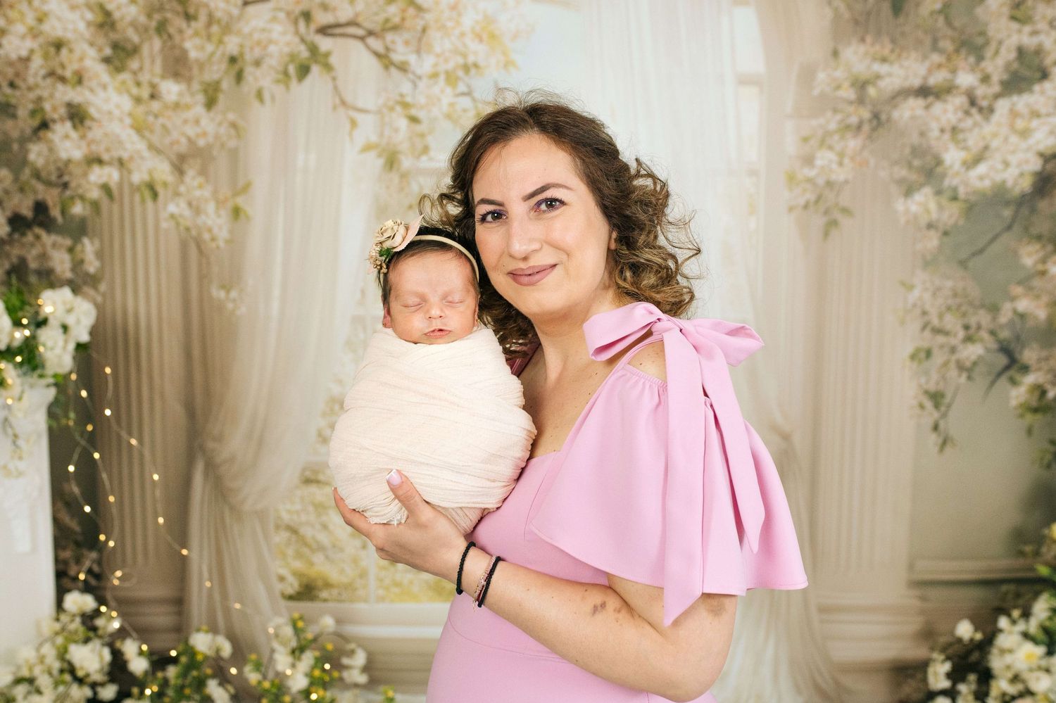 A woman in a pink dress holds a swaddled newborn baby with a floral headband in a white floral studio setting.