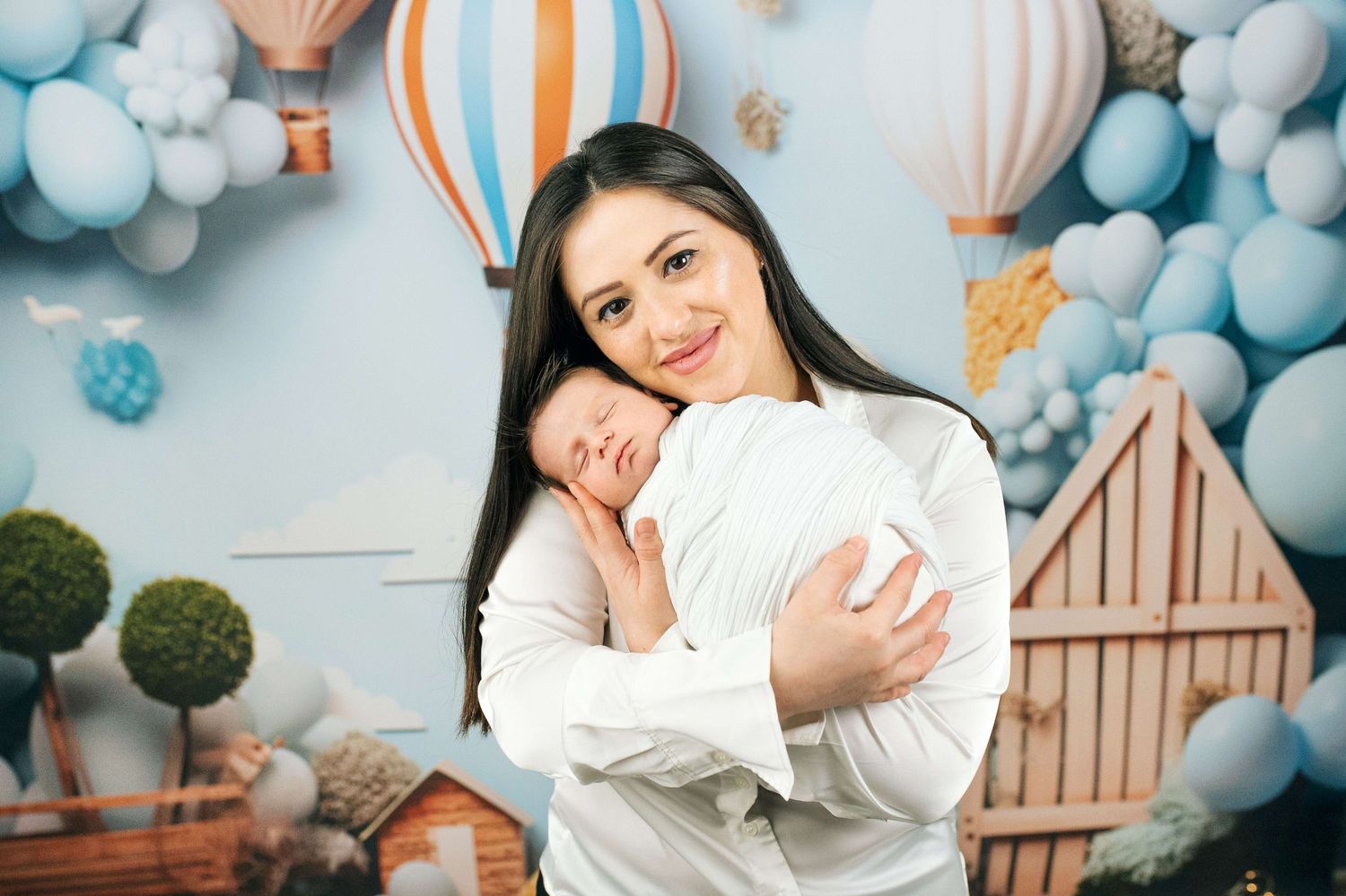 A young mother in a white shirt tenderly holds her sleeping newborn baby against a blue balloon nursery backdrop.