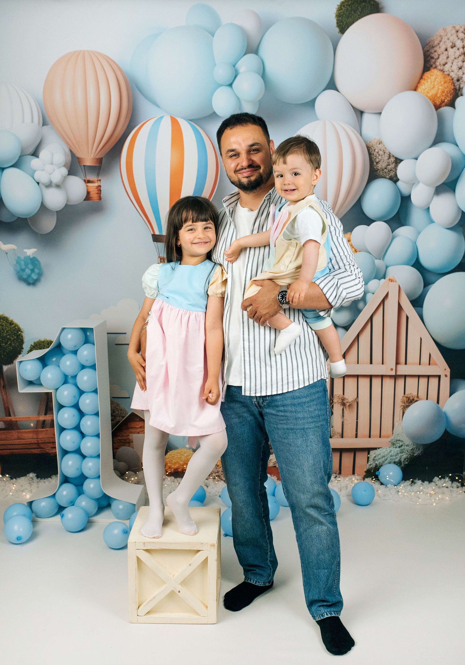 Father holds baby boy while daughter stands beside him at a first birthday party with blue balloon backdrop.
