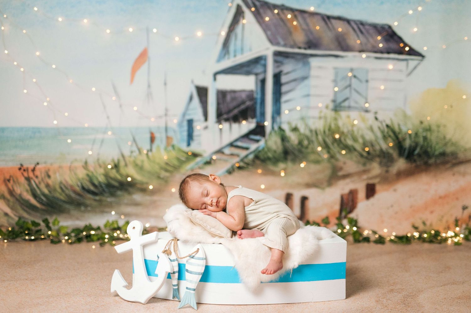Newborn baby sleeping peacefully in a decorative boat bed with a beach hut painted backdrop and twinkling fairy lights.