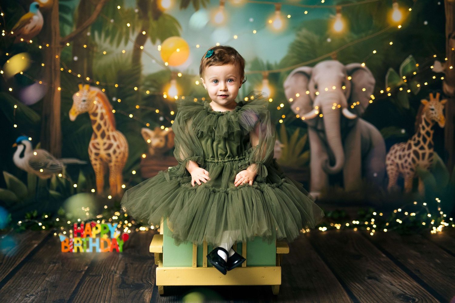 Toddler girl in green tutu dress sits on bench with jungle animal backdrop and fairy lights at birthday party.