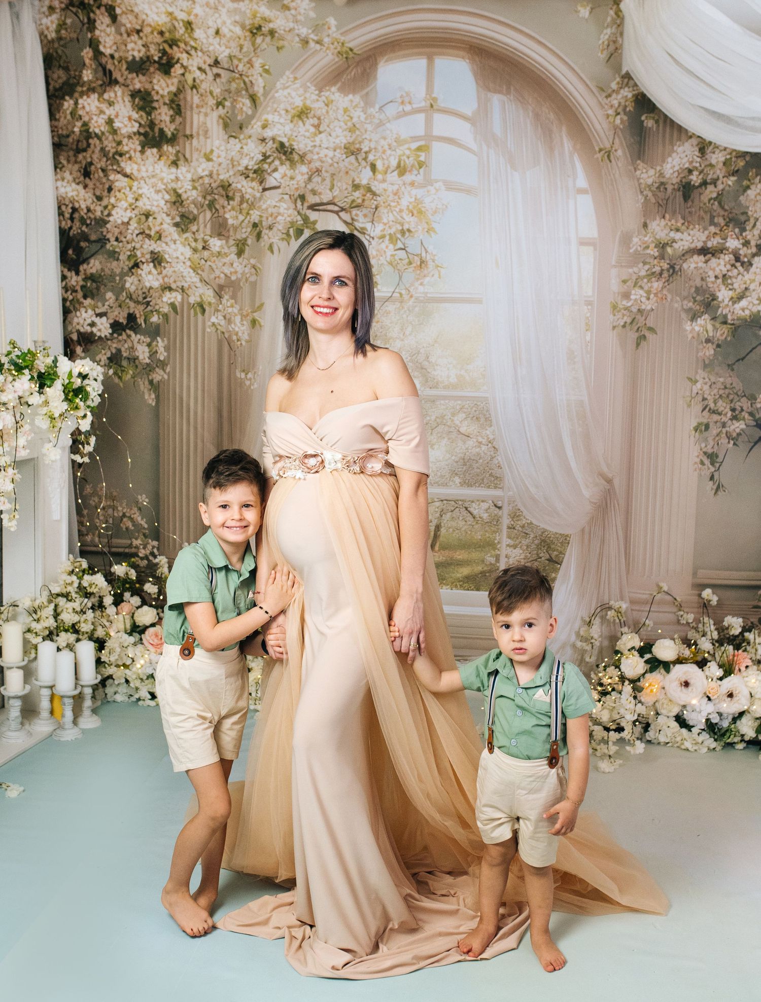 Pregnant woman in elegant beige gown poses with two young boys in green shirts amid floral studio backdrop.