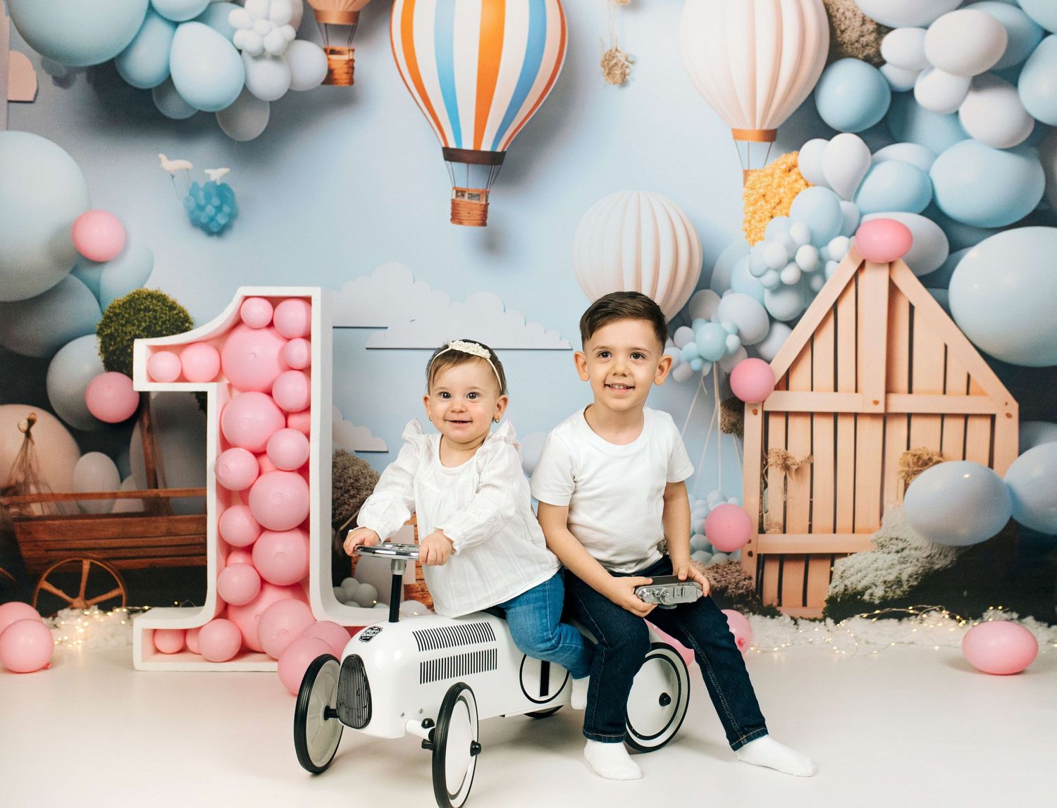 Two children pose with toy car and giant number 1 balloon backdrop for a first birthday photo session.