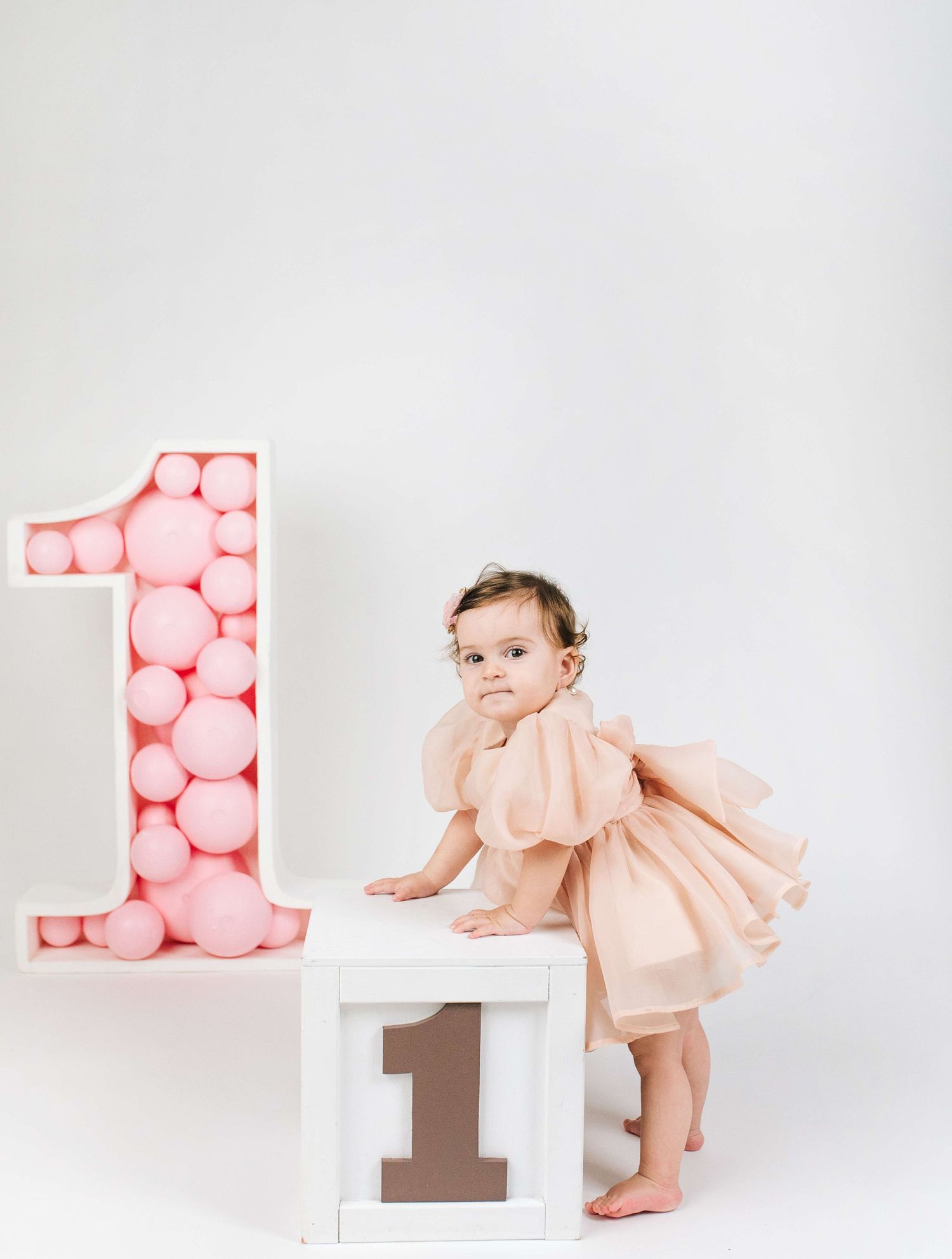 A baby girl in a peach dress poses beside a large pink balloon number one for her first birthday photo.