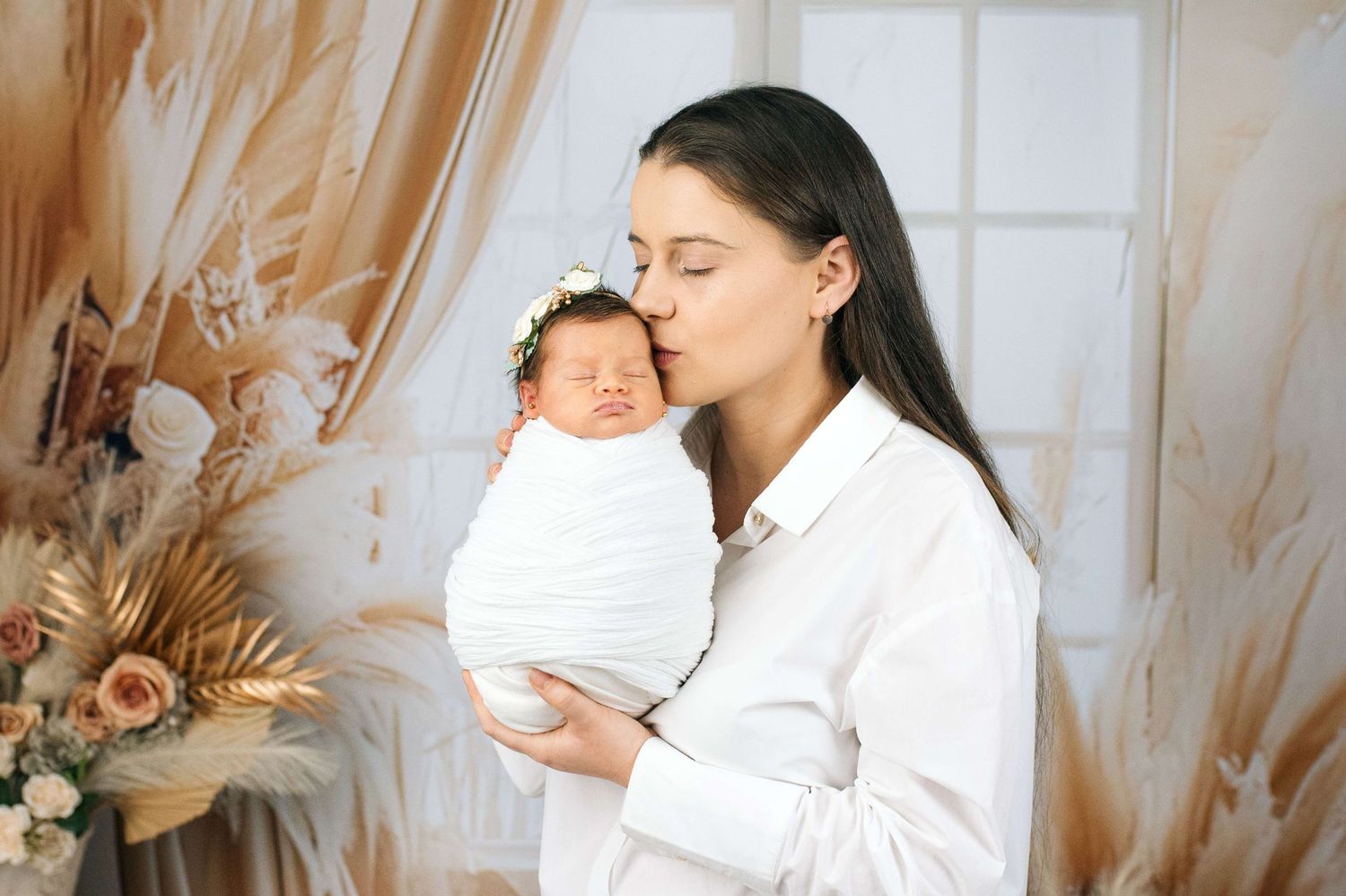 A young mother in a white shirt gently kisses her sleeping newborn wrapped in white, in a boho studio setting.