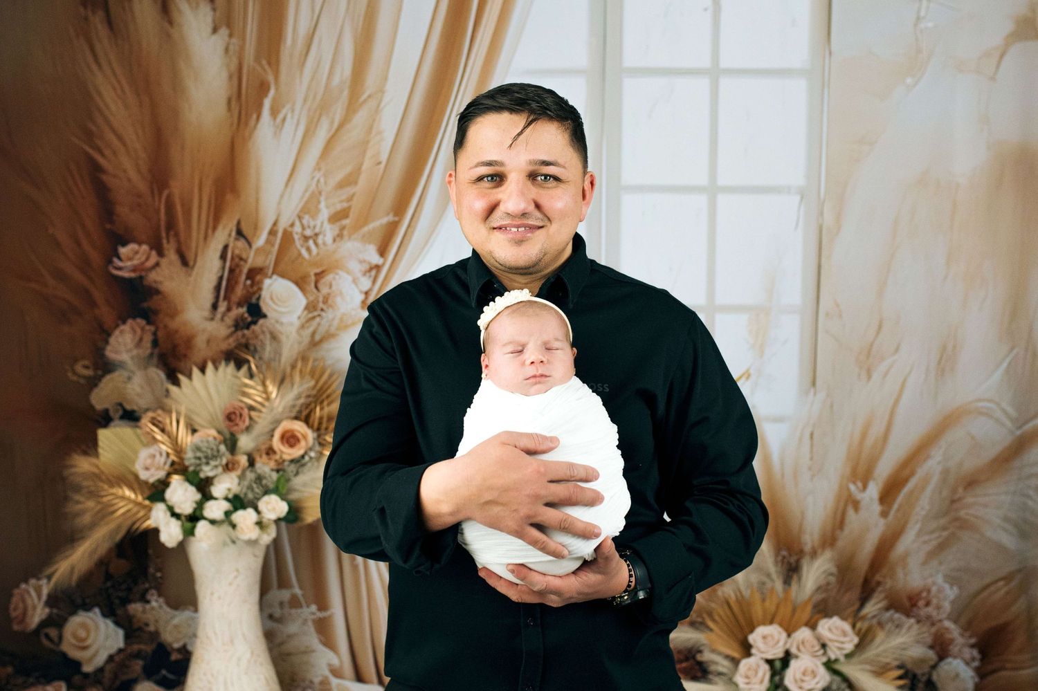 Man in black shirt proudly holds swaddled newborn baby during professional photo session with floral backdrop.