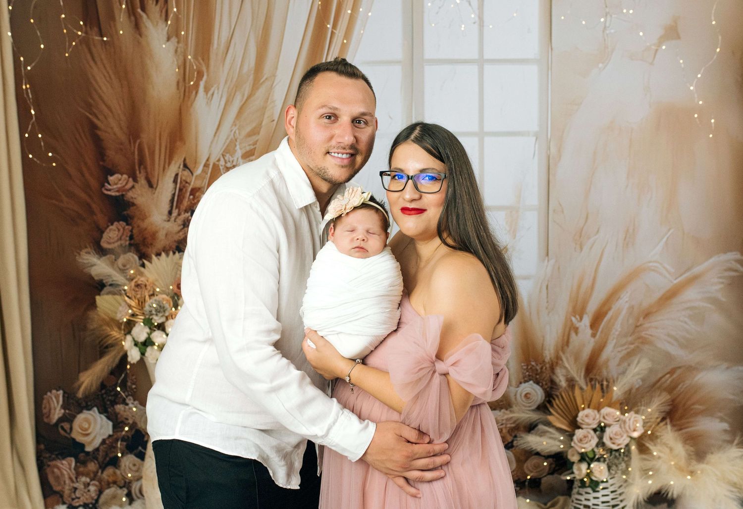 A happy couple in a floral studio setting proudly holds their newborn baby wrapped in white.