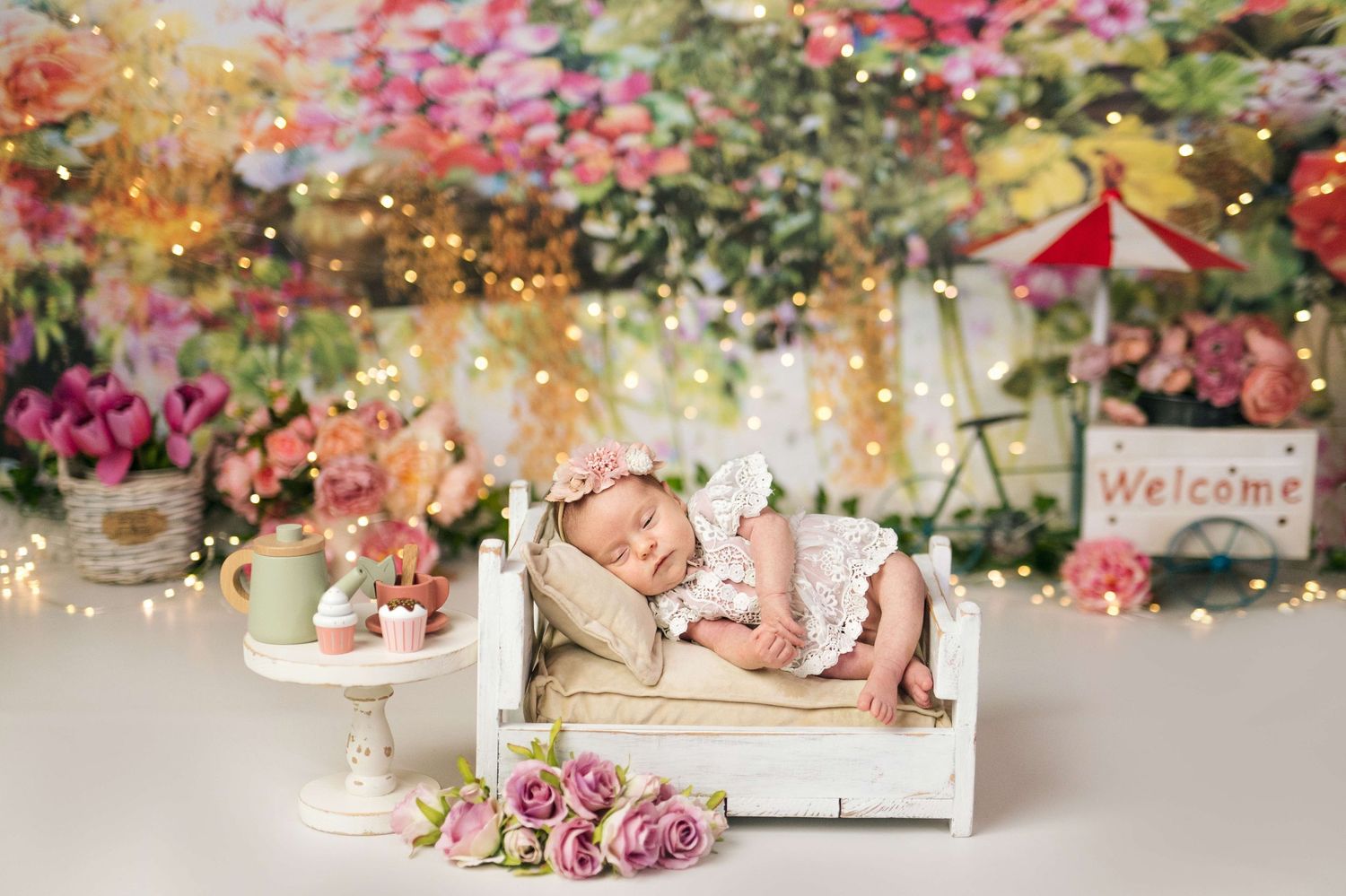 Sleeping newborn baby in white lace outfit on miniature bench surrounded by pink roses and floral backdrop with fairy lights.