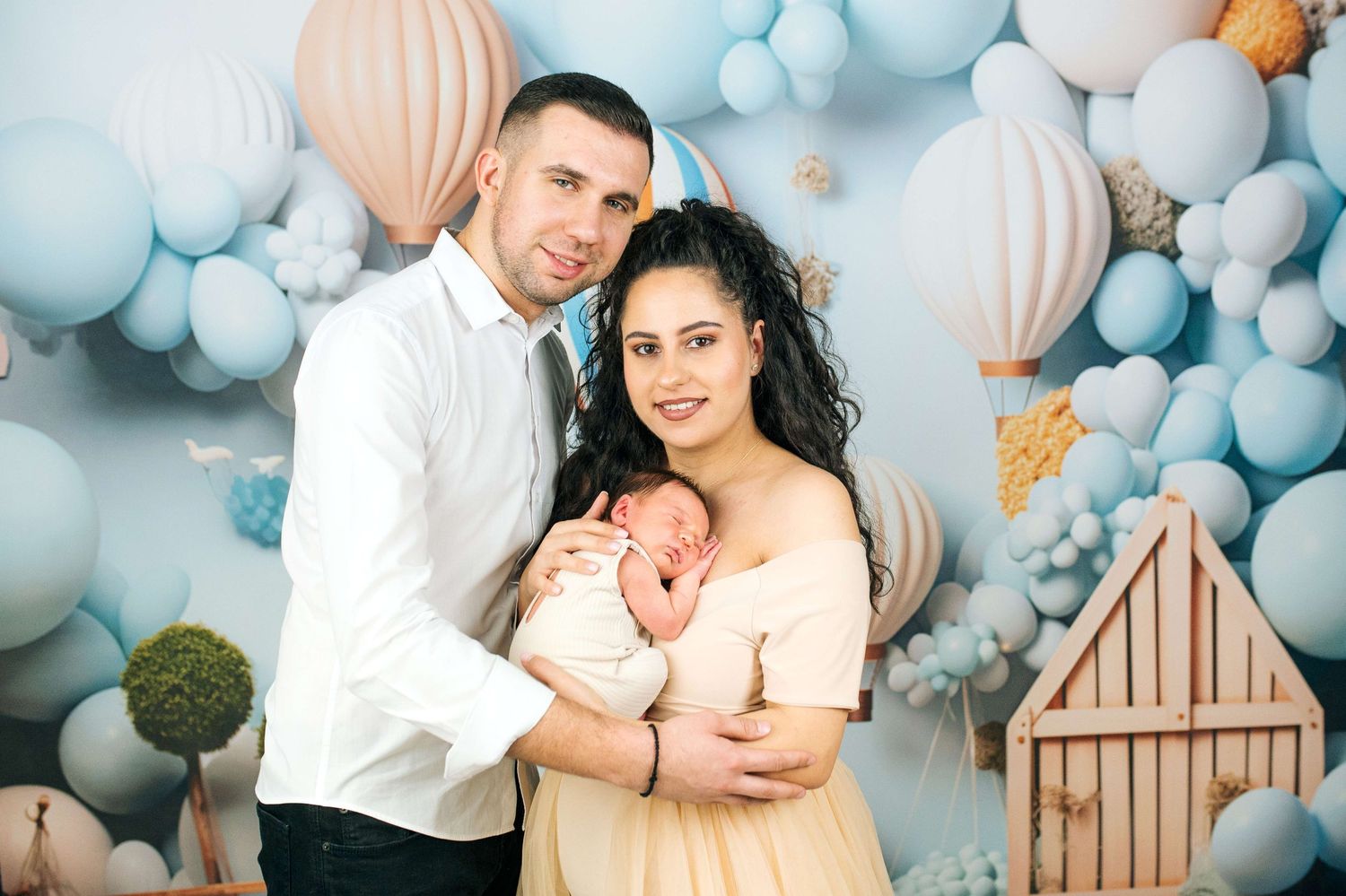 Young couple holding newborn baby in front of blue balloon backdrop for professional family photo shoot.