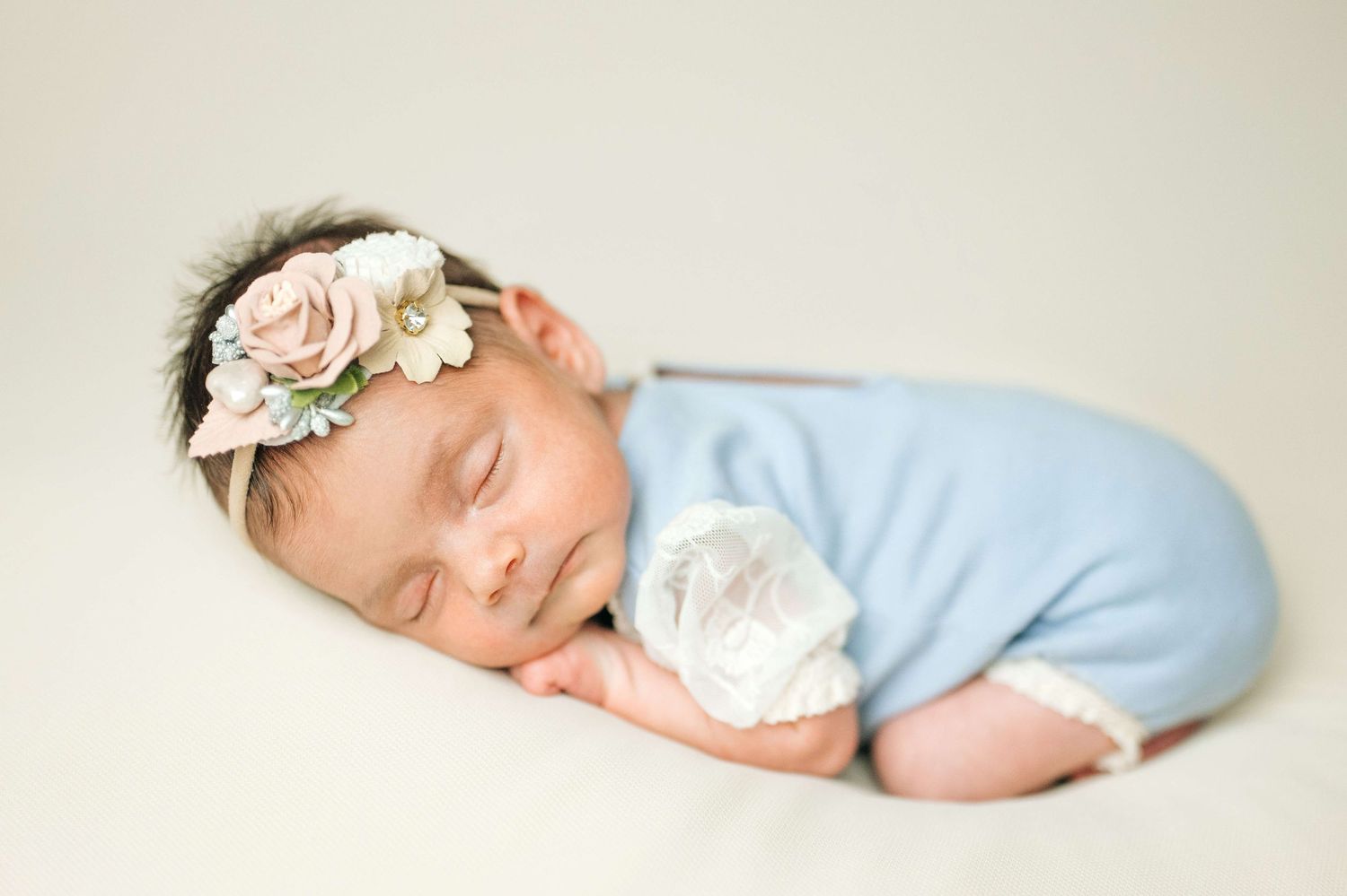 Sleeping newborn baby girl in blue romper and floral headband posed on cream backdrop.