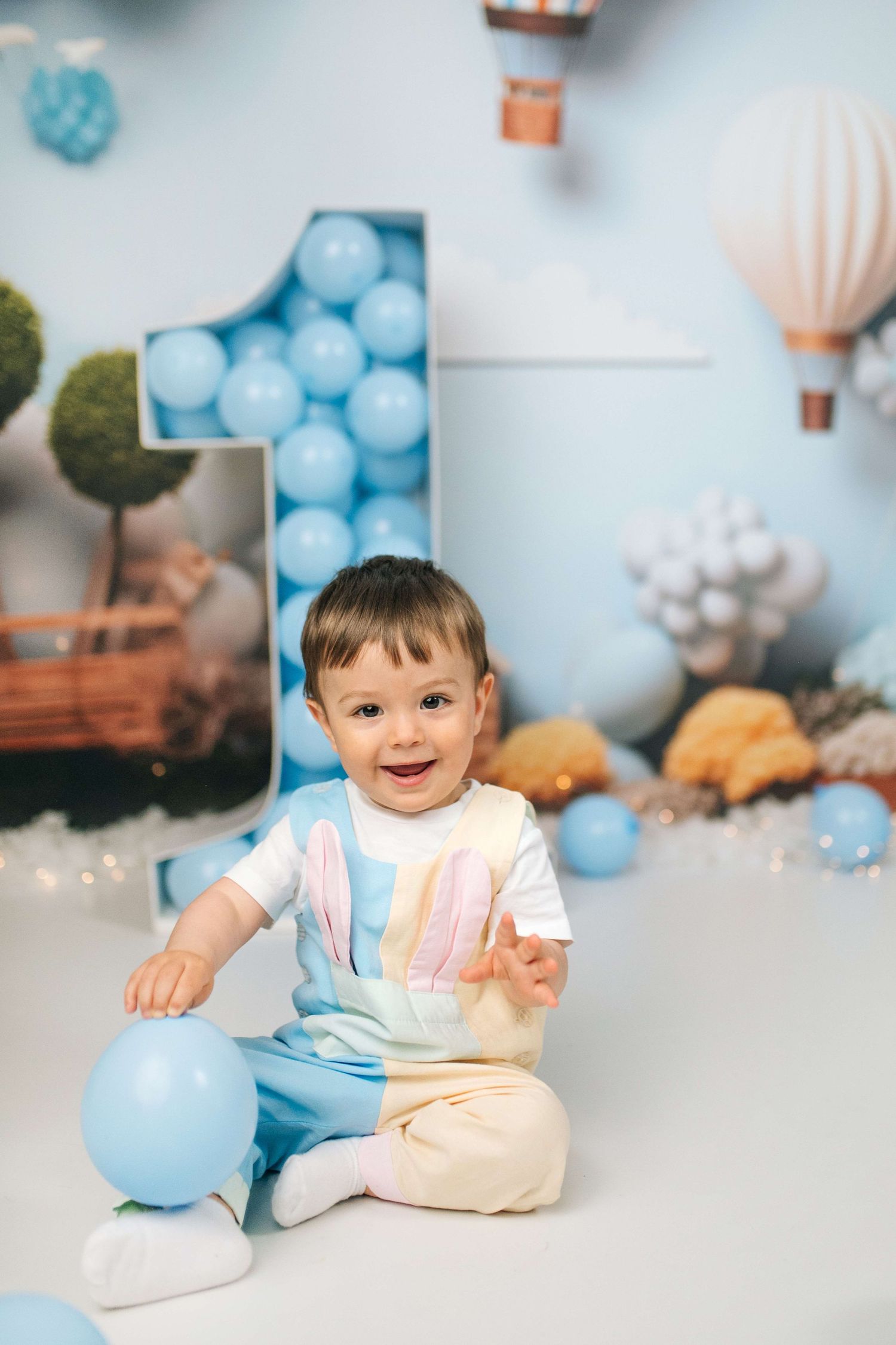 Smiling baby boy sitting on floor at first birthday party with blue balloons and number one decoration backdrop.