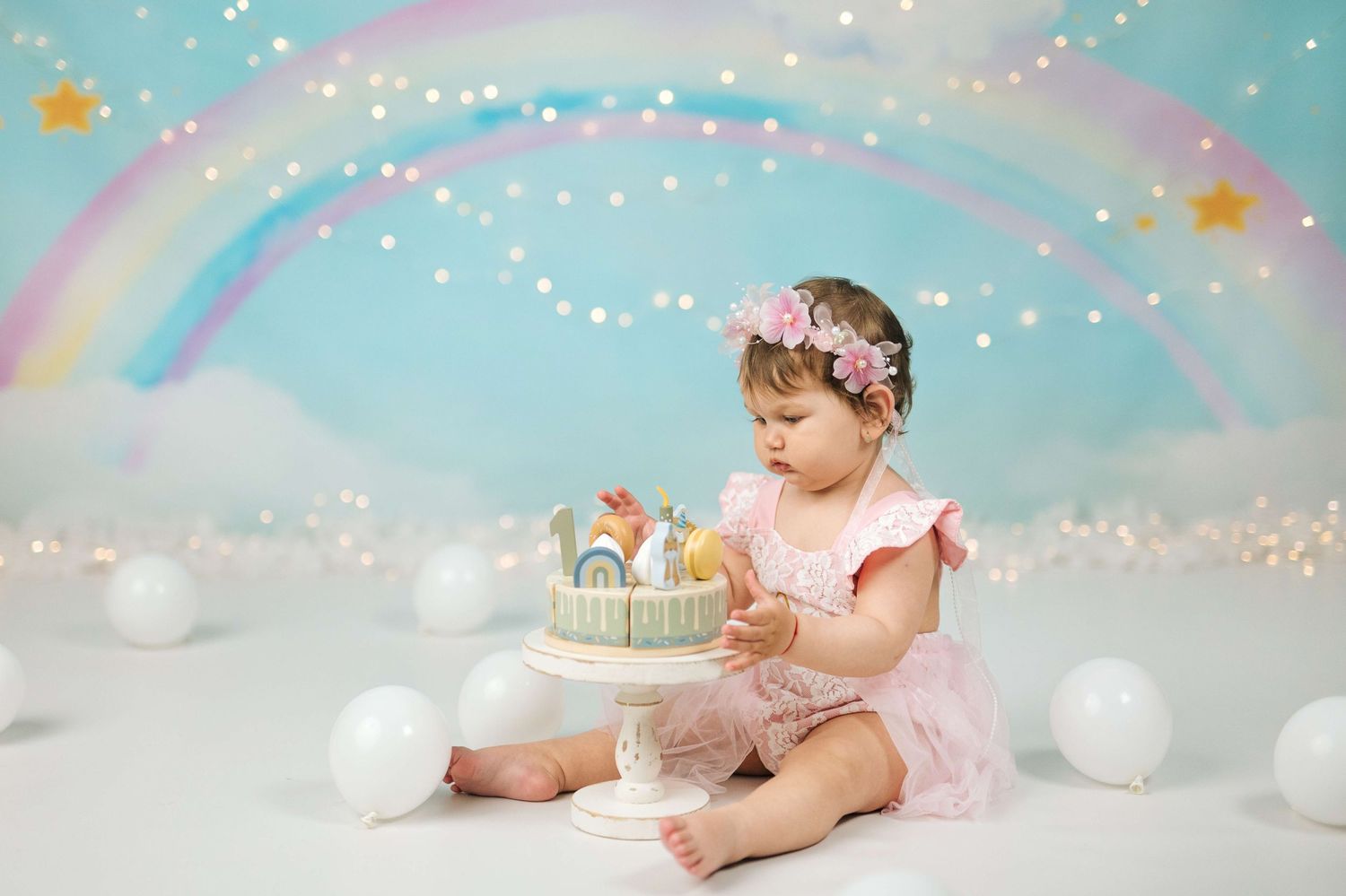 Baby girl in pink tutu sits with rainbow birthday cake, balloons, and fairy lights backdrop for first birthday cake smash.