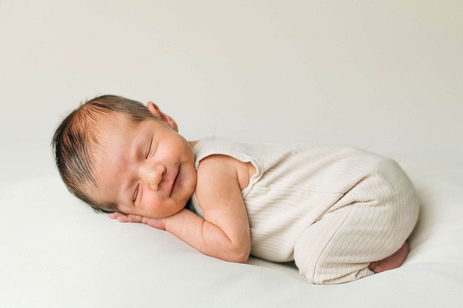 Smiling newborn baby in cream romper sleeping peacefully on white backdrop in professional photo.