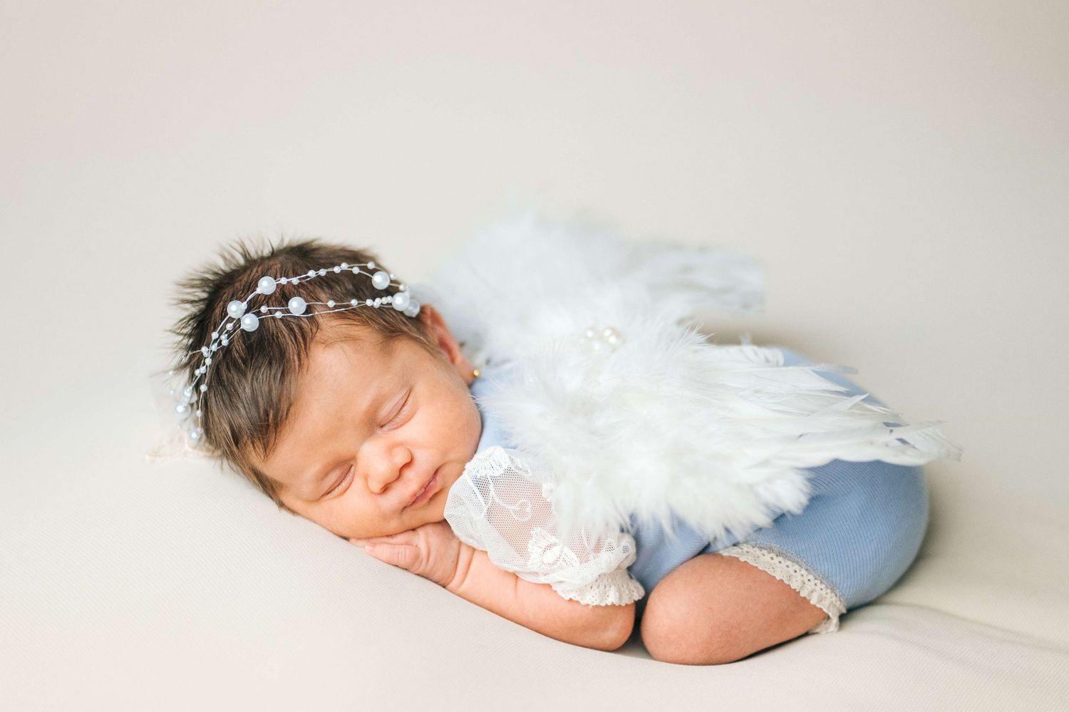 Sleeping newborn baby wearing white feather angel wings and floral headband on a soft beige backdrop.