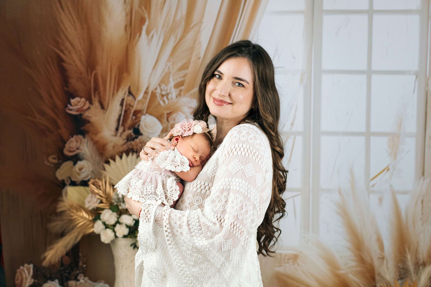 Young woman with long dark hair in white sweater holds sleeping newborn baby in boho-styled photo studio.
