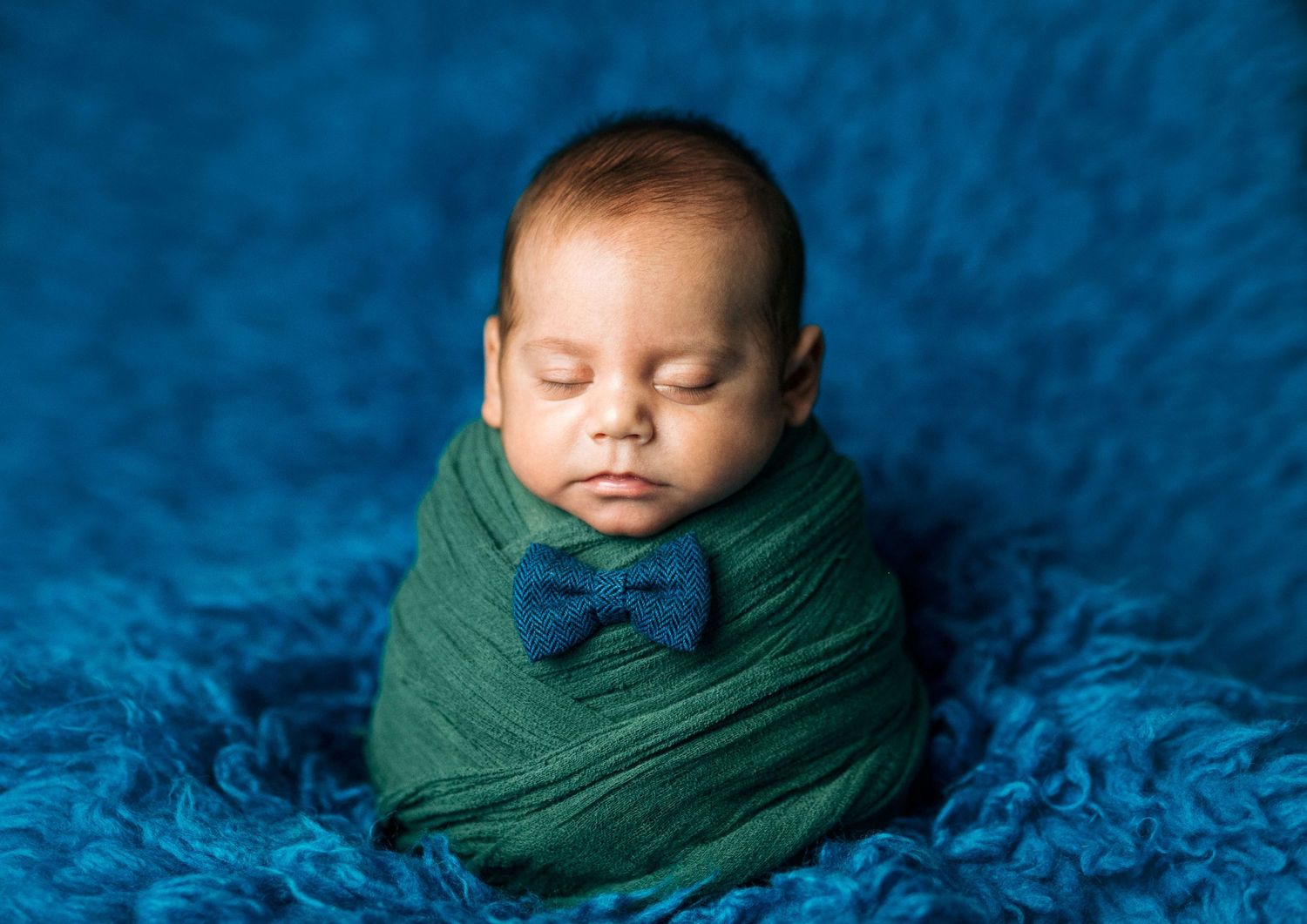 Sleeping newborn baby swaddled in green wrap with blue bow tie on blue fluffy blanket.