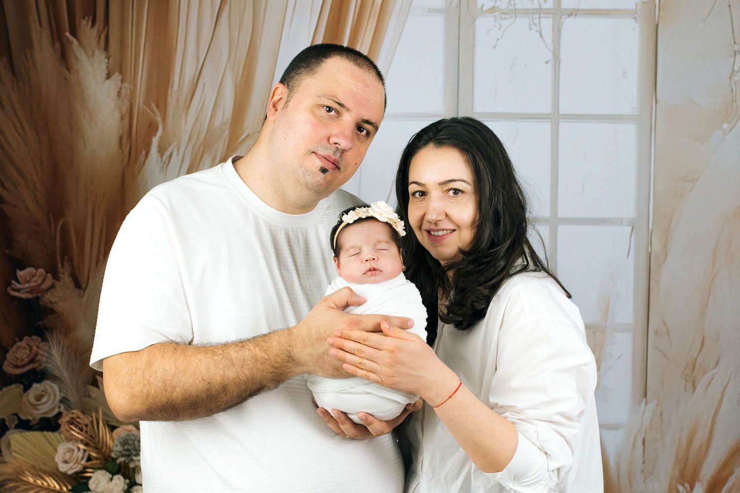 Happy couple holding their newborn baby wrapped in white, posing for a family portrait indoors.