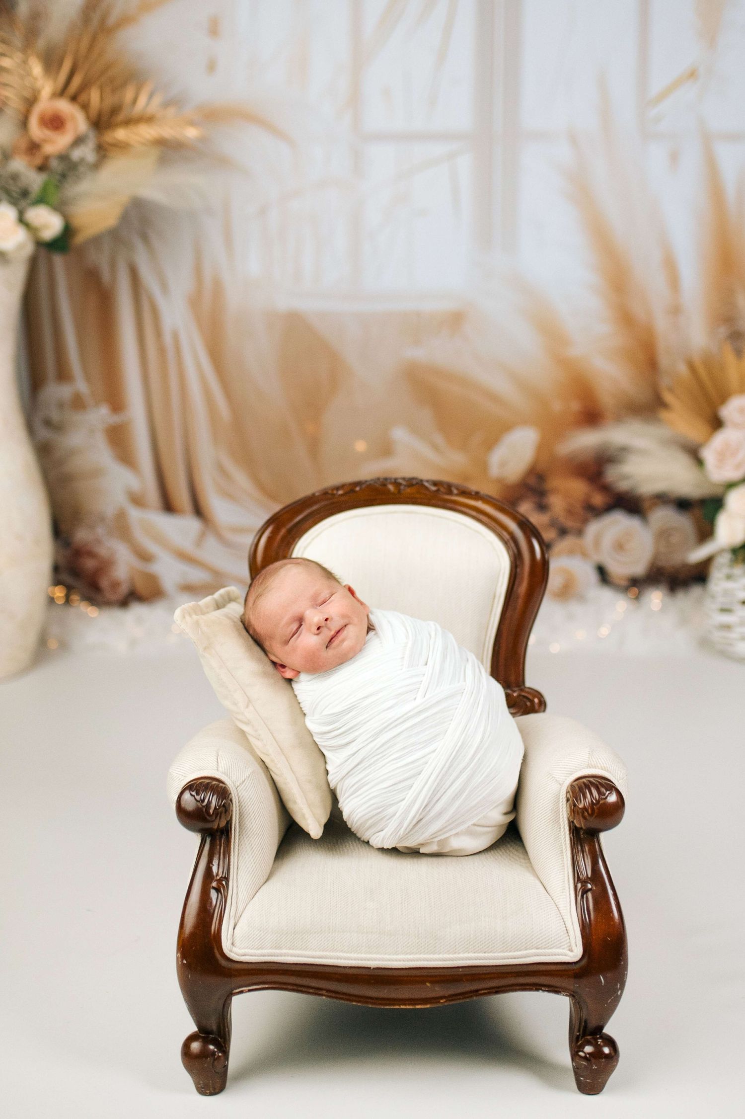Newborn baby swaddled in white wrap sleeping peacefully on a miniature cream armchair in a boho studio setting.