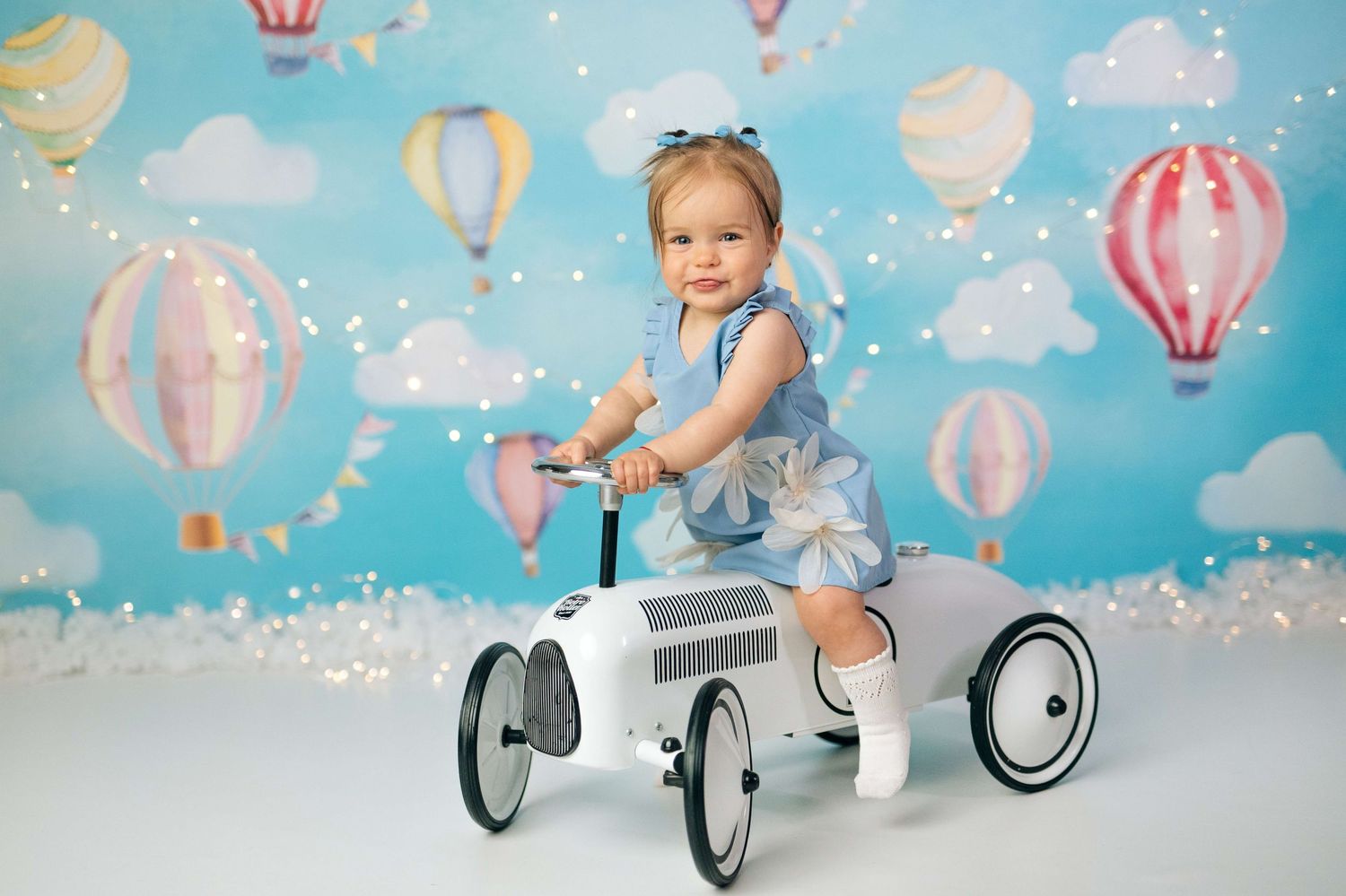Toddler girl in blue dress riding a white toy car against a colorful hot air balloon backdrop with twinkling lights.