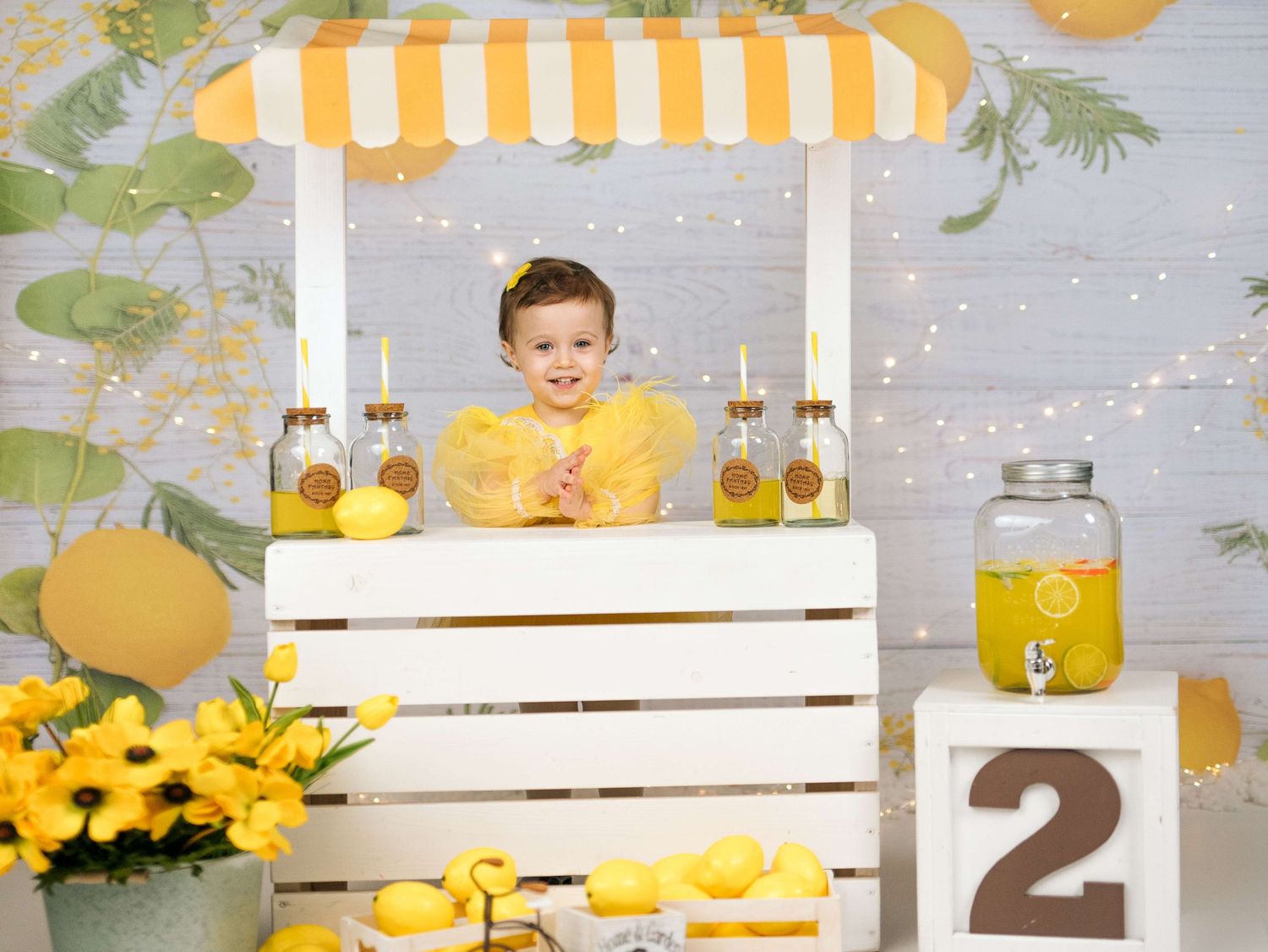 Toddler in yellow outfit smiling at a white lemonade stand with sunflowers, lemons, and a number 2 block.