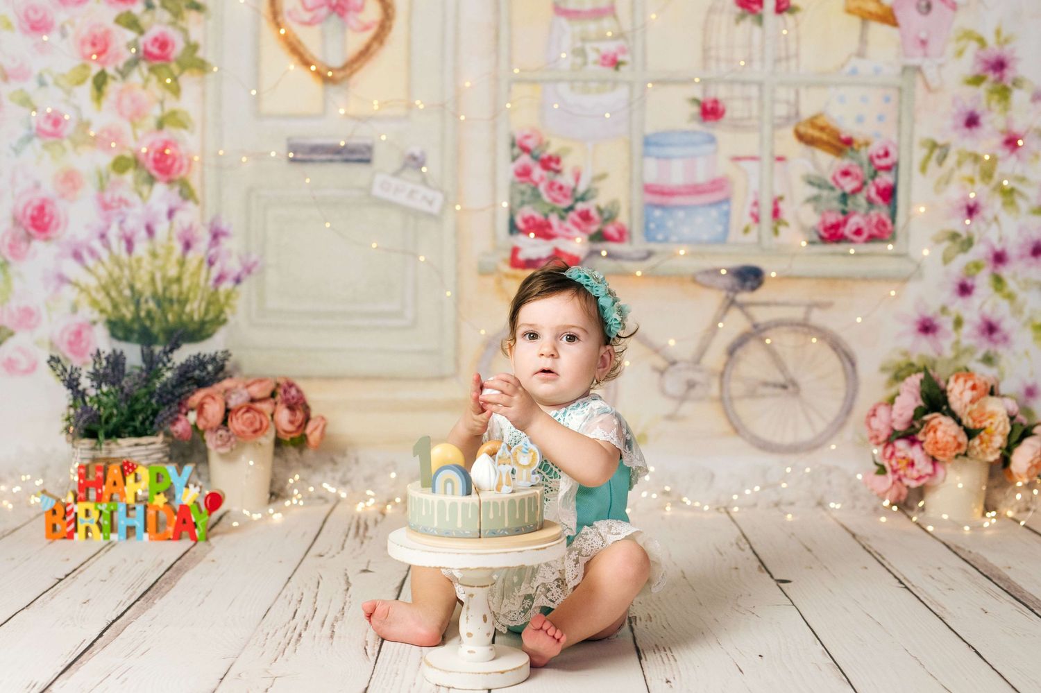Baby girl celebrates first birthday with cake smash session against floral backdrop with fairy lights.