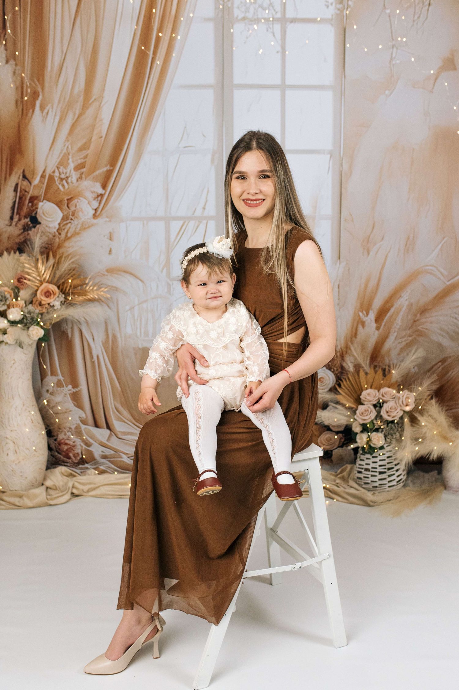 Young woman in brown dress holds baby girl in white outfit, posed in elegant studio with floral decor.