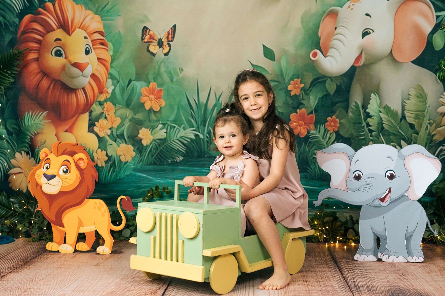 Two children pose with a toy jeep against a colorful jungle safari backdrop with lions and elephants.