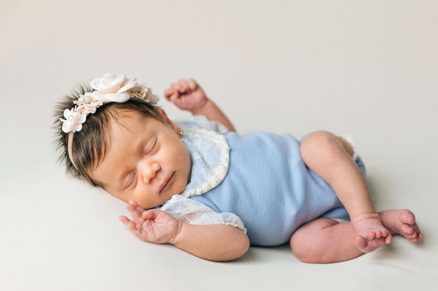 Sleeping newborn baby girl in blue romper and floral headband posed on white backdrop.