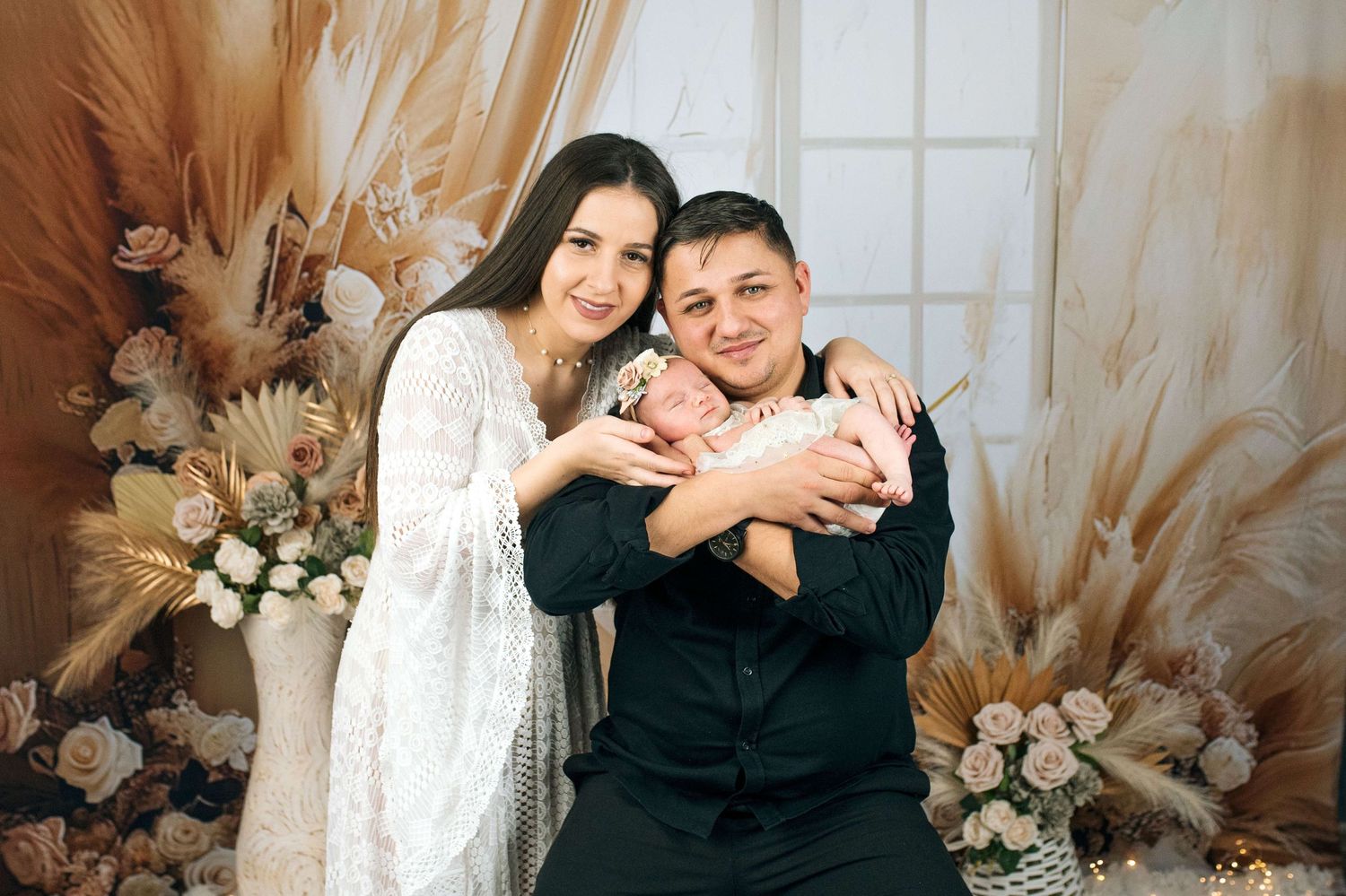 A couple in a boho-styled studio holding their newborn baby, surrounded by dried floral arrangements.