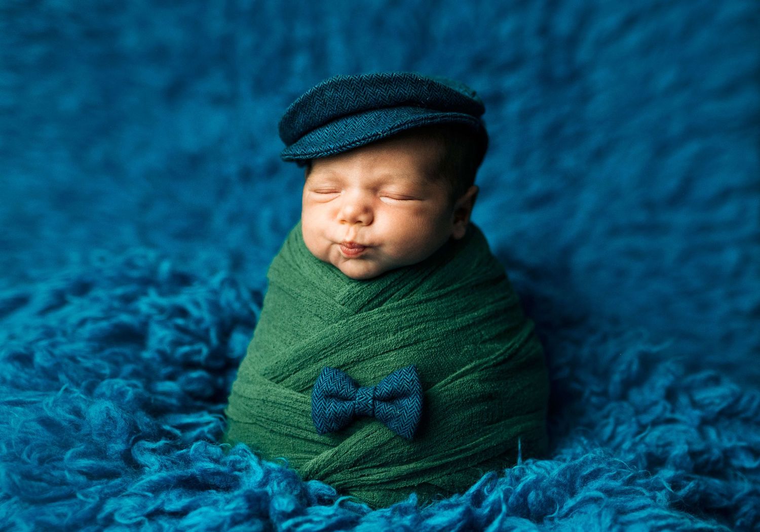 Sleeping newborn baby wrapped in green swaddle wearing blue bow tie and flat cap on blue textured background.