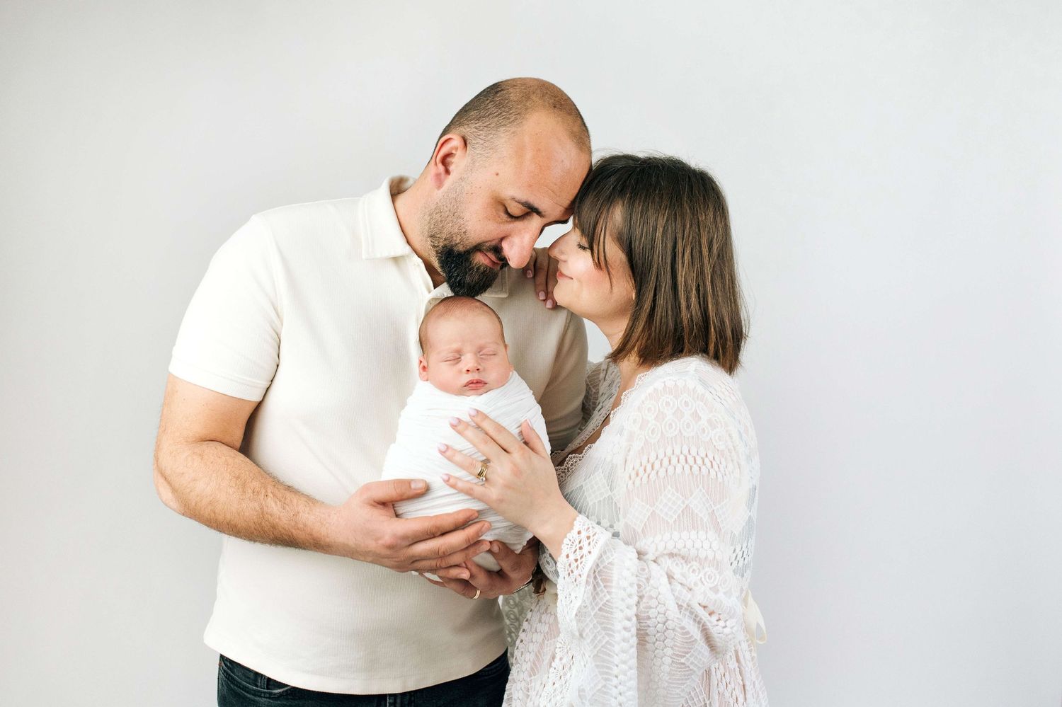 Happy parents with beards tenderly hold their newborn baby wrapped in white against a light background.