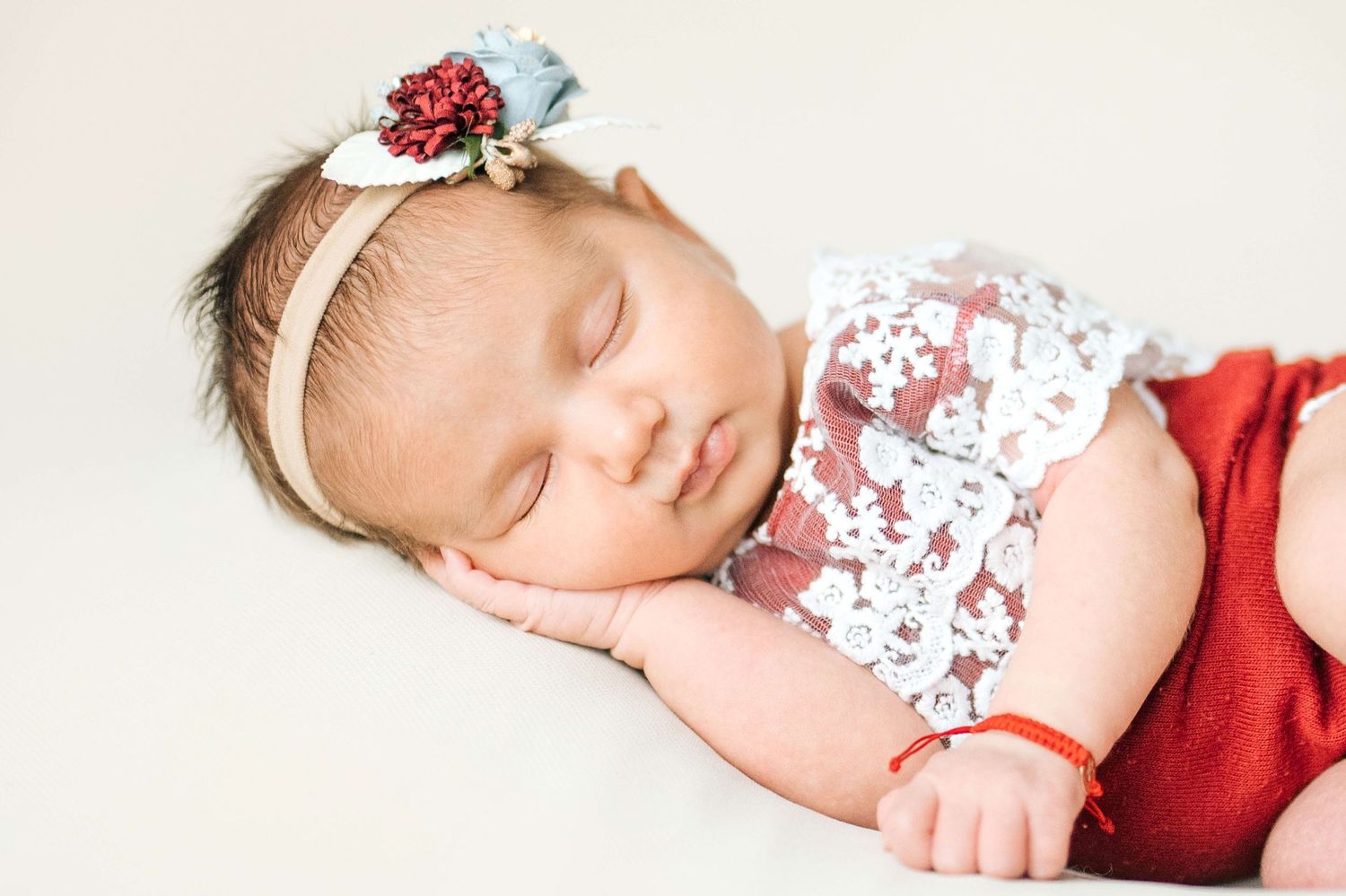 Sleeping newborn baby girl wearing a floral headband and red outfit with lace top in a professional portrait.
