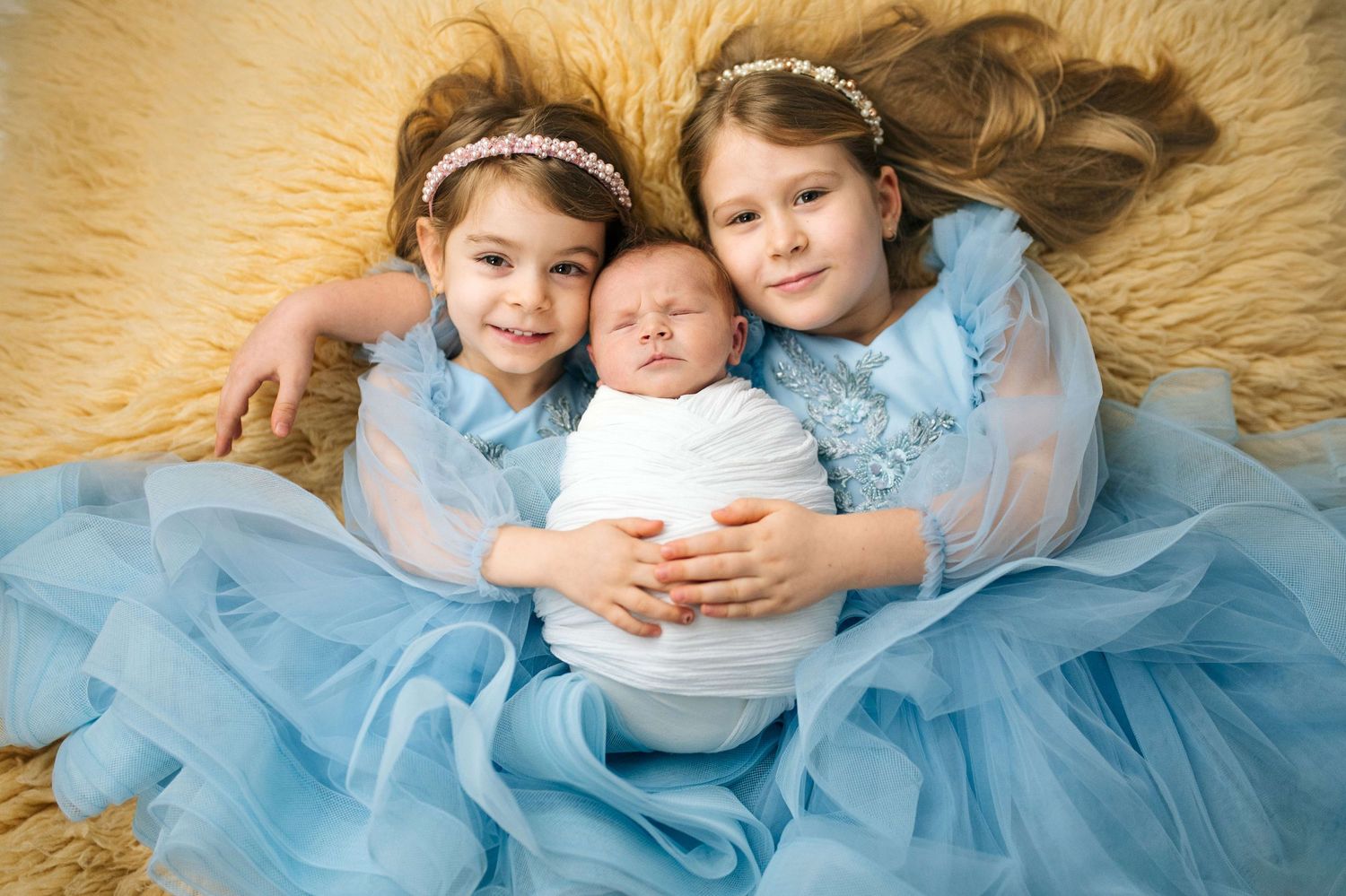 Two young girls in blue tulle dresses holding a swaddled newborn baby, posing on a fluffy beige backdrop.