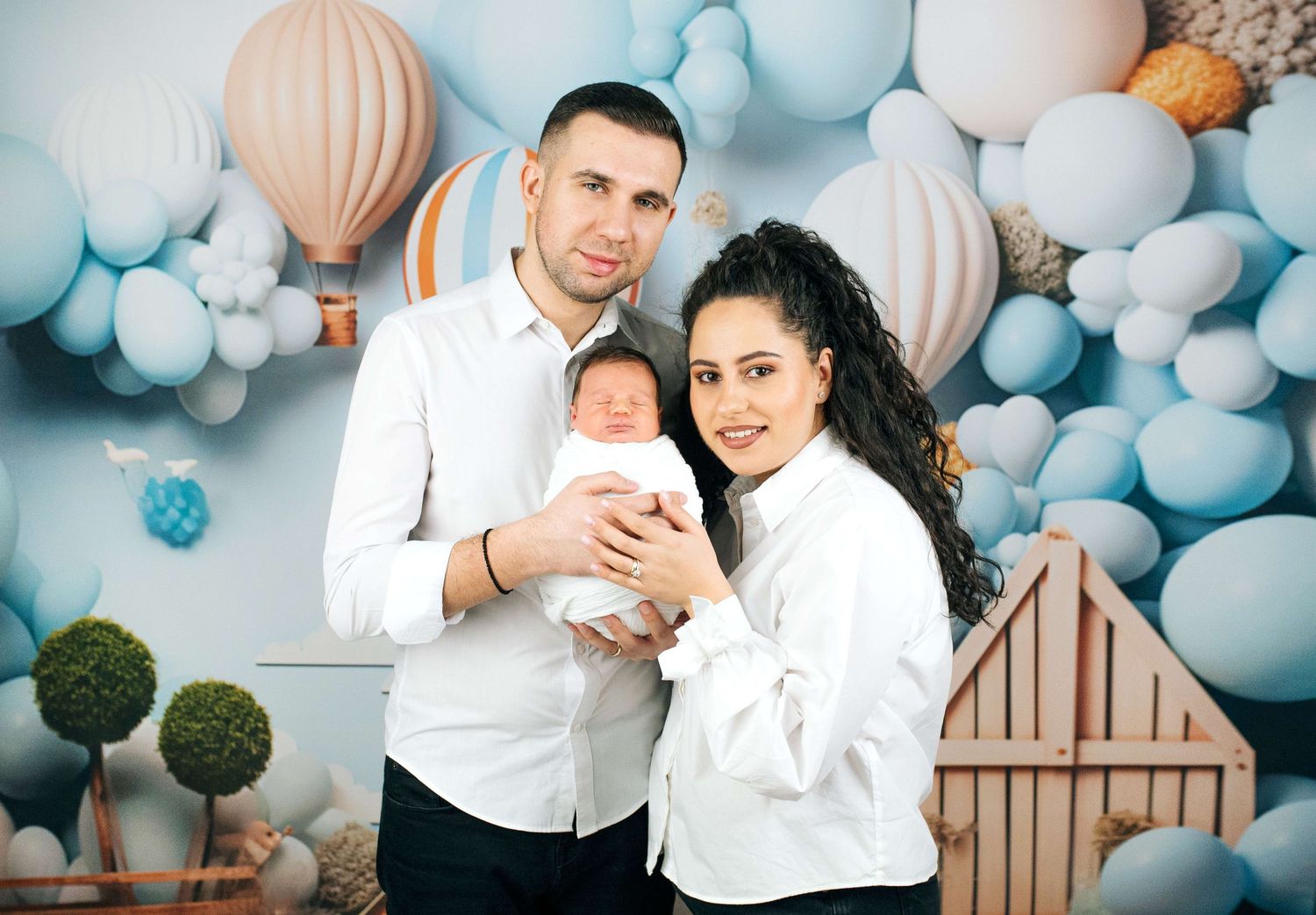 Happy couple holding newborn baby in front of blue balloon and hot air balloon themed backdrop.