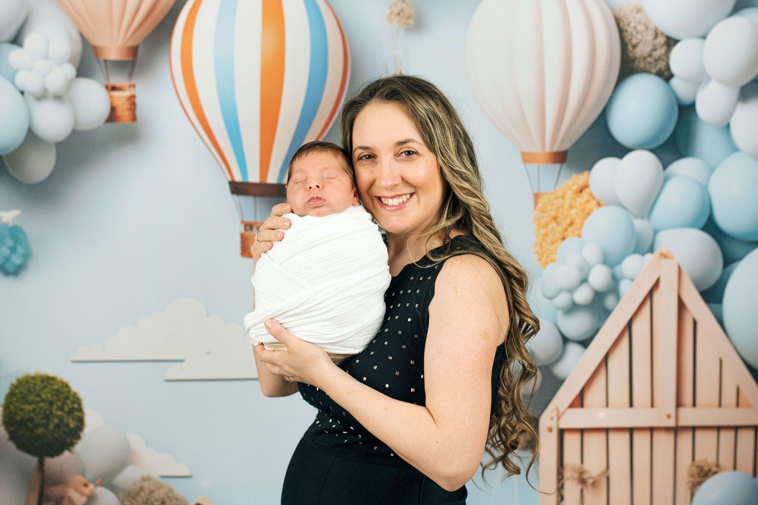 Smiling woman holding swaddled newborn baby in front of hot air balloon themed backdrop with pastel balloons.