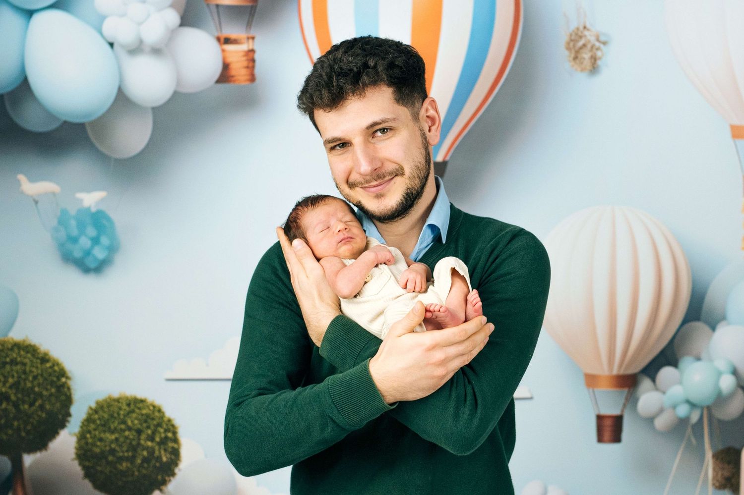 A smiling man in a green sweater holds a sleeping newborn baby against a blue balloon and hot air balloon backdrop.
