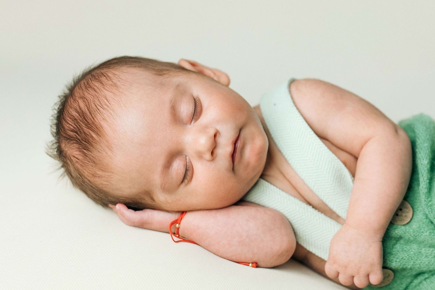 Peaceful newborn baby sleeping on stomach wearing green wrap with red bracelet on wrist against light background.