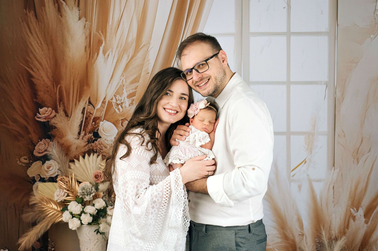 Happy couple in white outfits holding newborn baby, posing in front of boho floral backdrop with pampas grass.